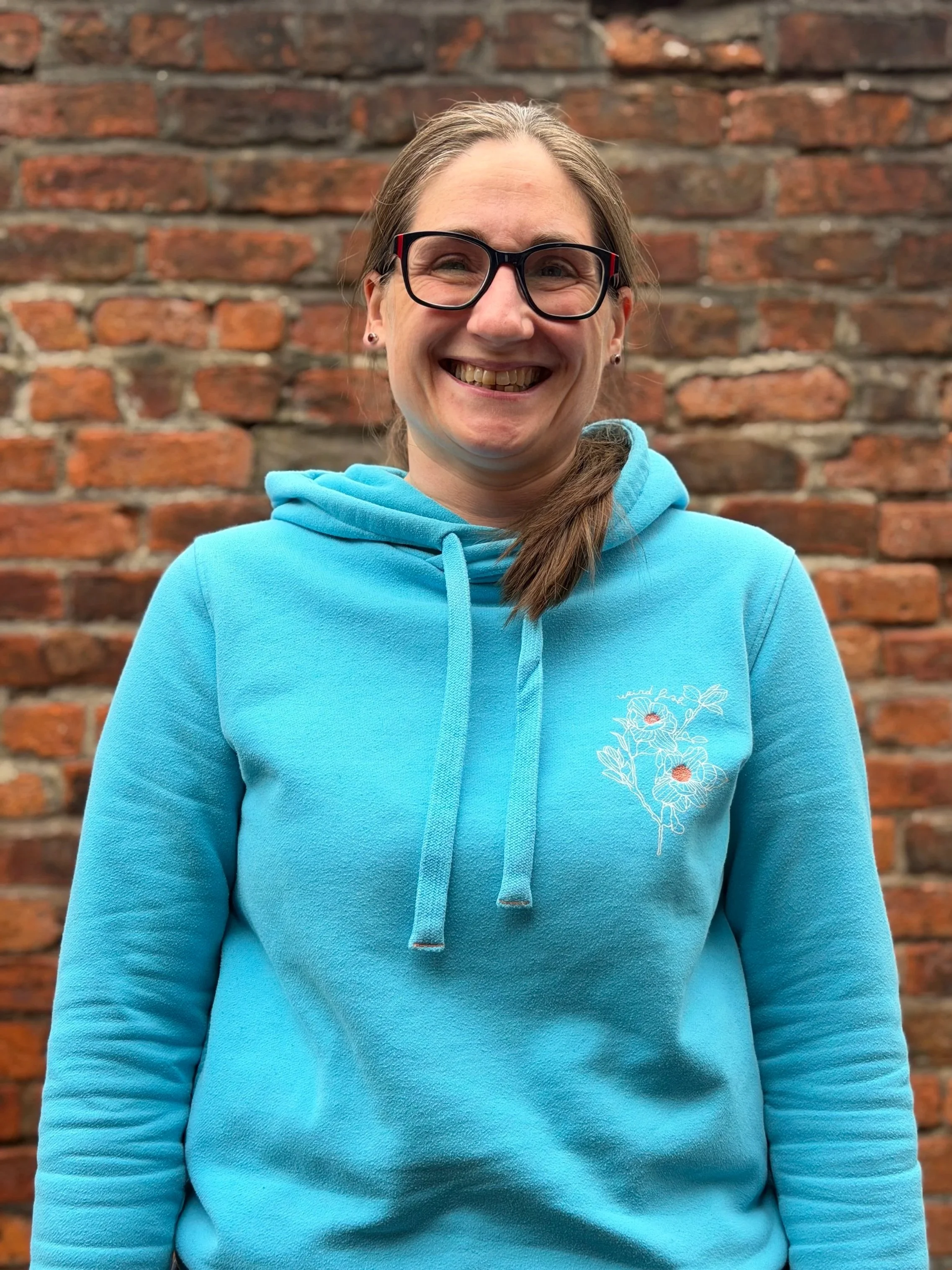 Rachel - A woman with long brown hair and glasses smiling at the camera, wearing a blue hoodie with a floral design on the chest, standing in front of a brick wall.