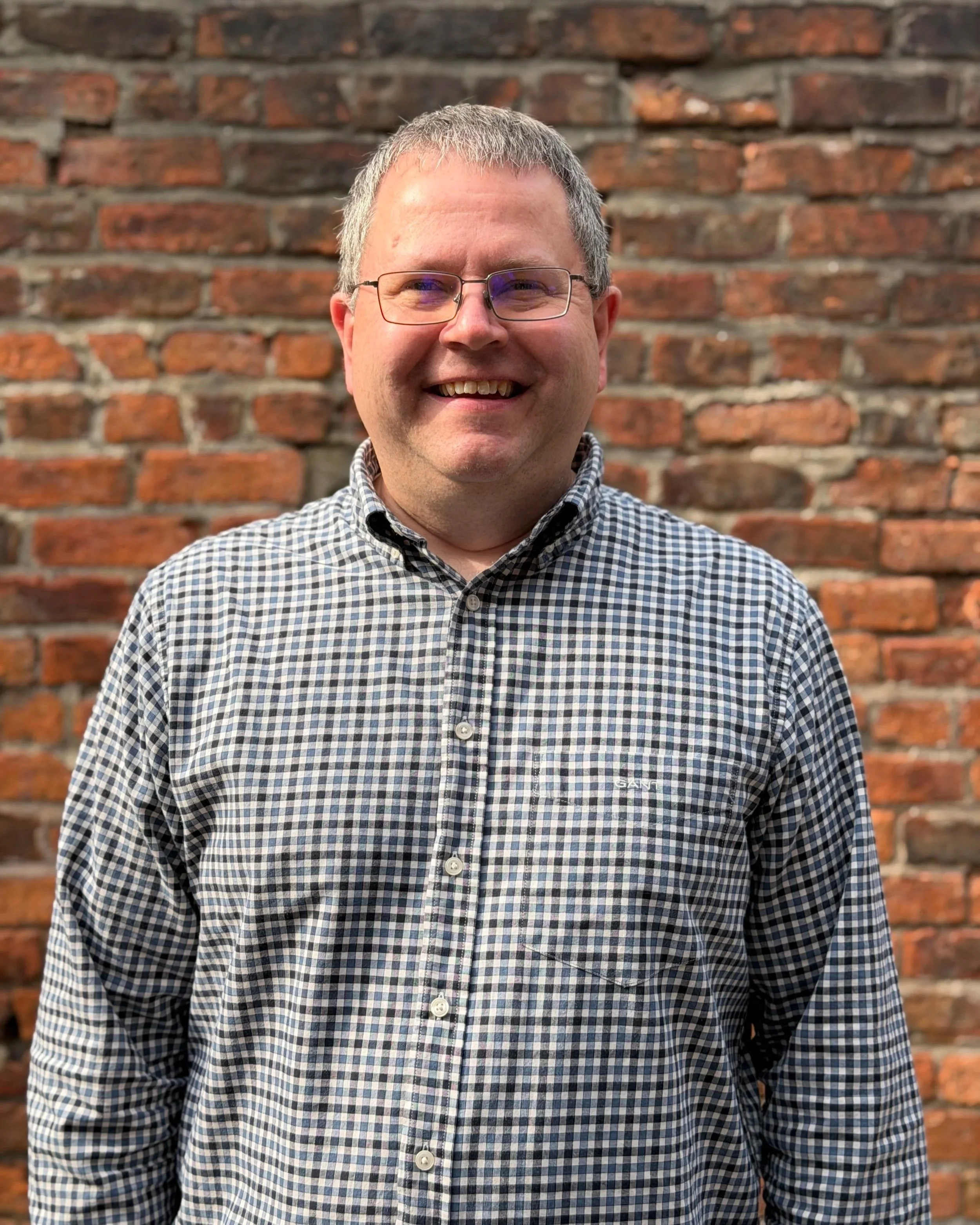 Ross - A smiling man with short hair and glasses wearing a checkered shirt standing in front of a brick wall.