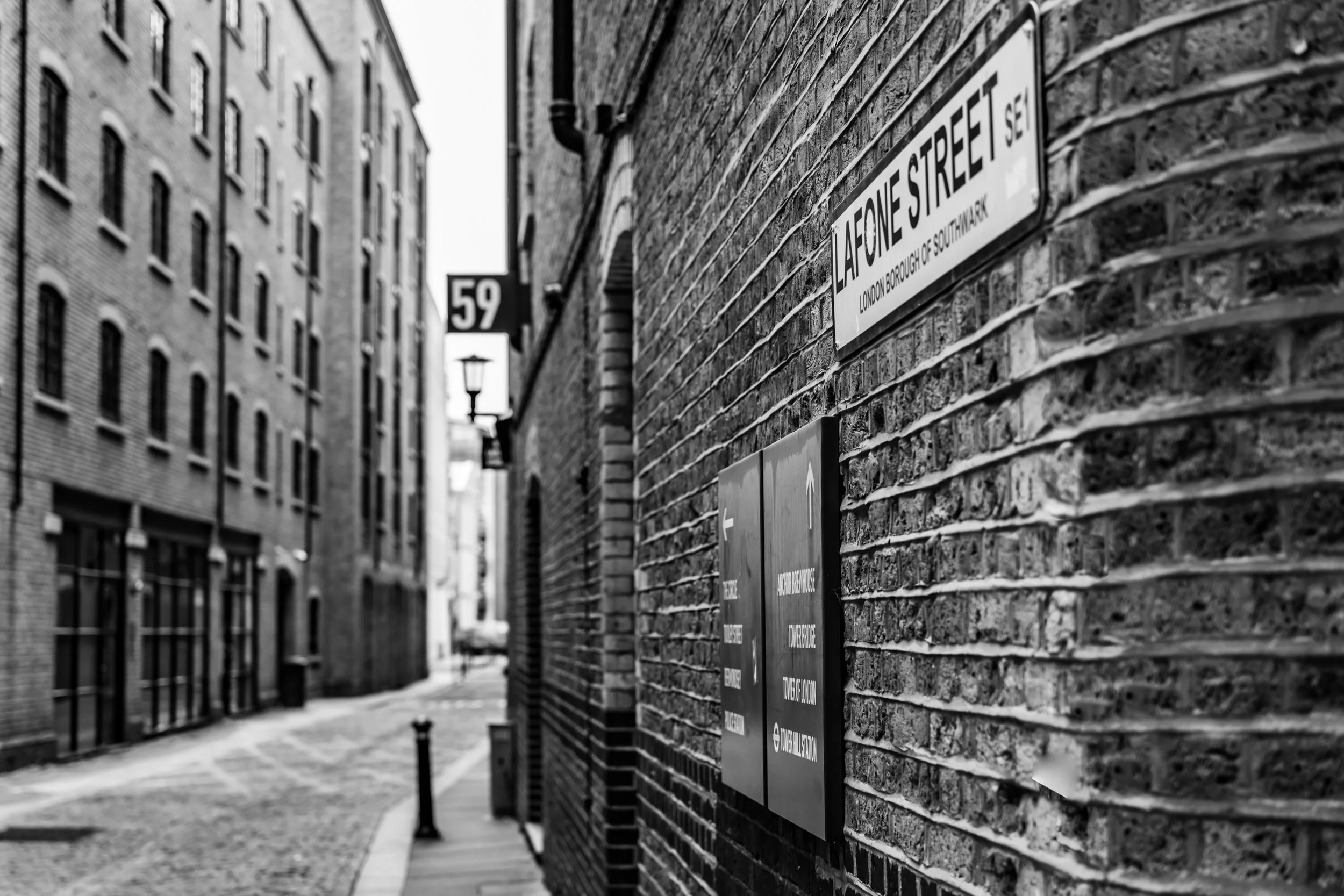 Black and white photo of a narrow street in London with brick buildings and street signs on the wall reading "La Fone Street" and "59".