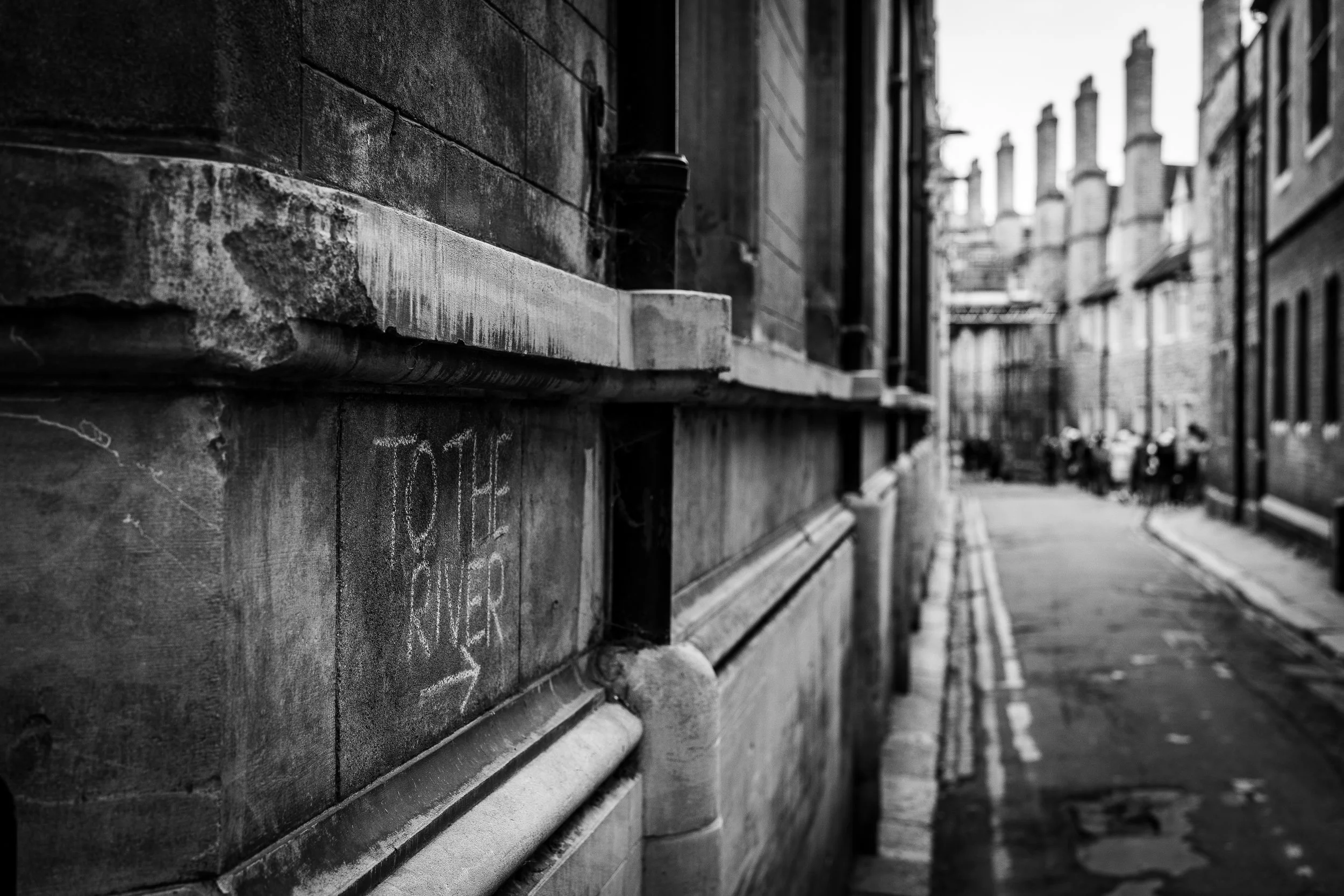 Black and white photo of an urban street scene with old brick and stone buildings, graffiti on the wall, and a row of chimneys above the rooflines. The street is narrow with a double yellow line and some blurred pedestrians in the distance.