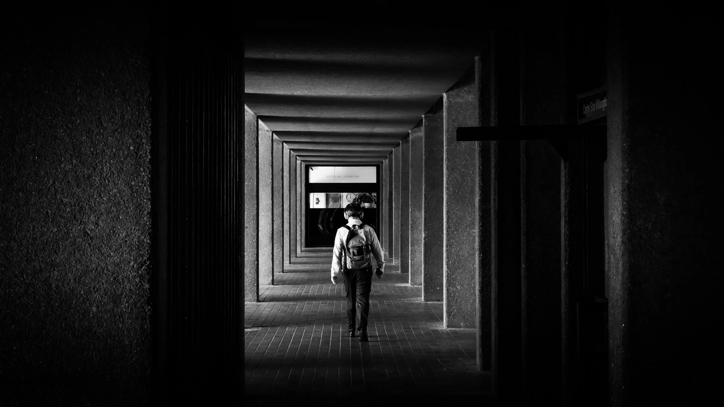 A person with a backpack walking through a long, dark corridor with repeating rectangular openings, in black and white.