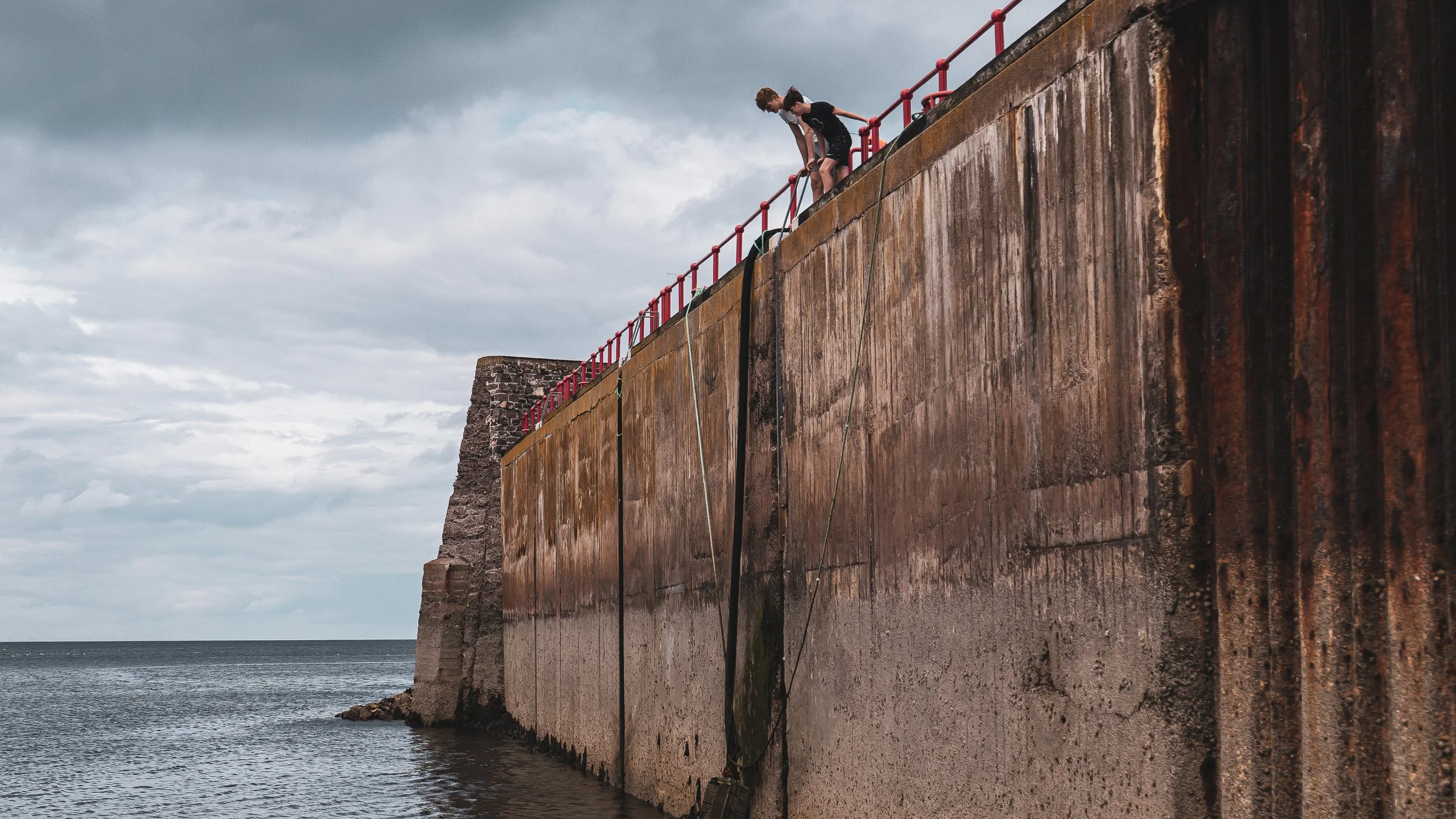Two boys climbing on a rusty seawall along the ocean under cloudy sky.