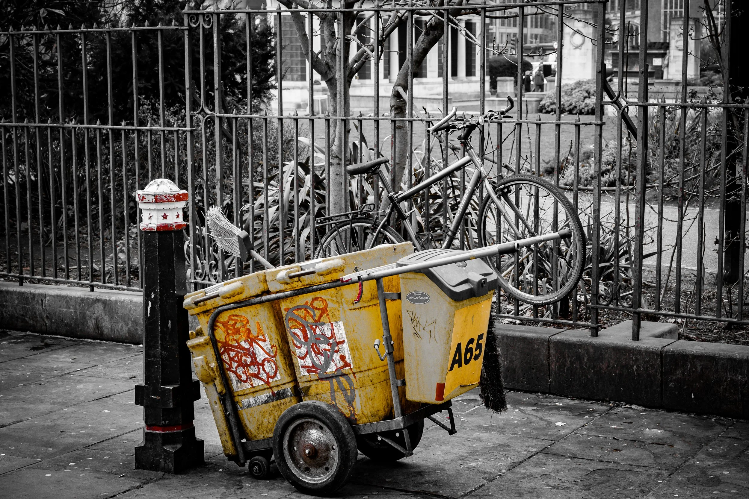 A yellow garbage bin filled with graffiti, placed next to a metal fence, with a bicycle hanging on the fence behind it. In the background, a tree and some buildings are visible, with a few people walking.