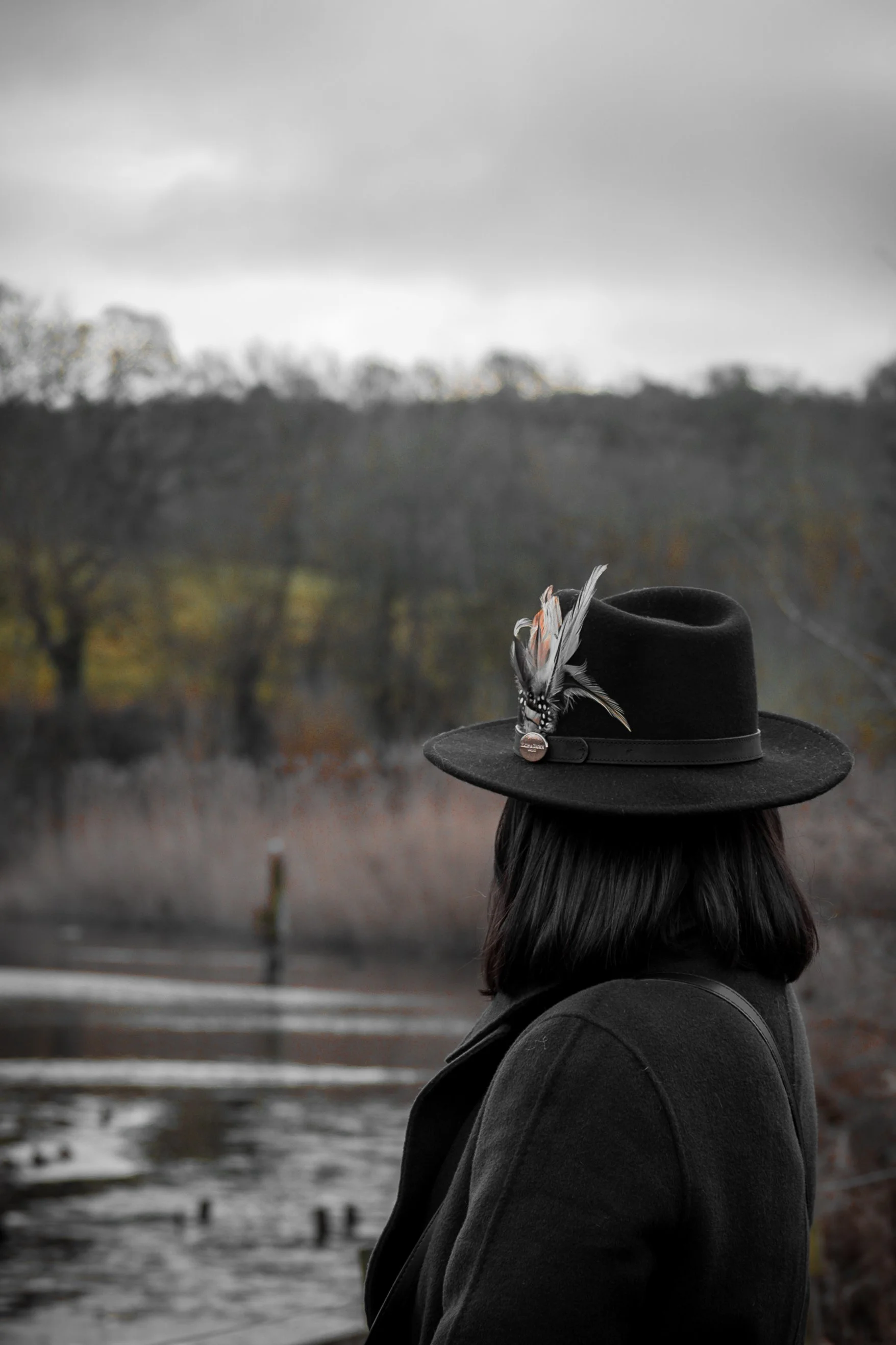 A woman with dark hair, wearing a dark coat and a black hat with a feather decoration, standing outdoors near a body of water with a forest in the background on an overcast day.