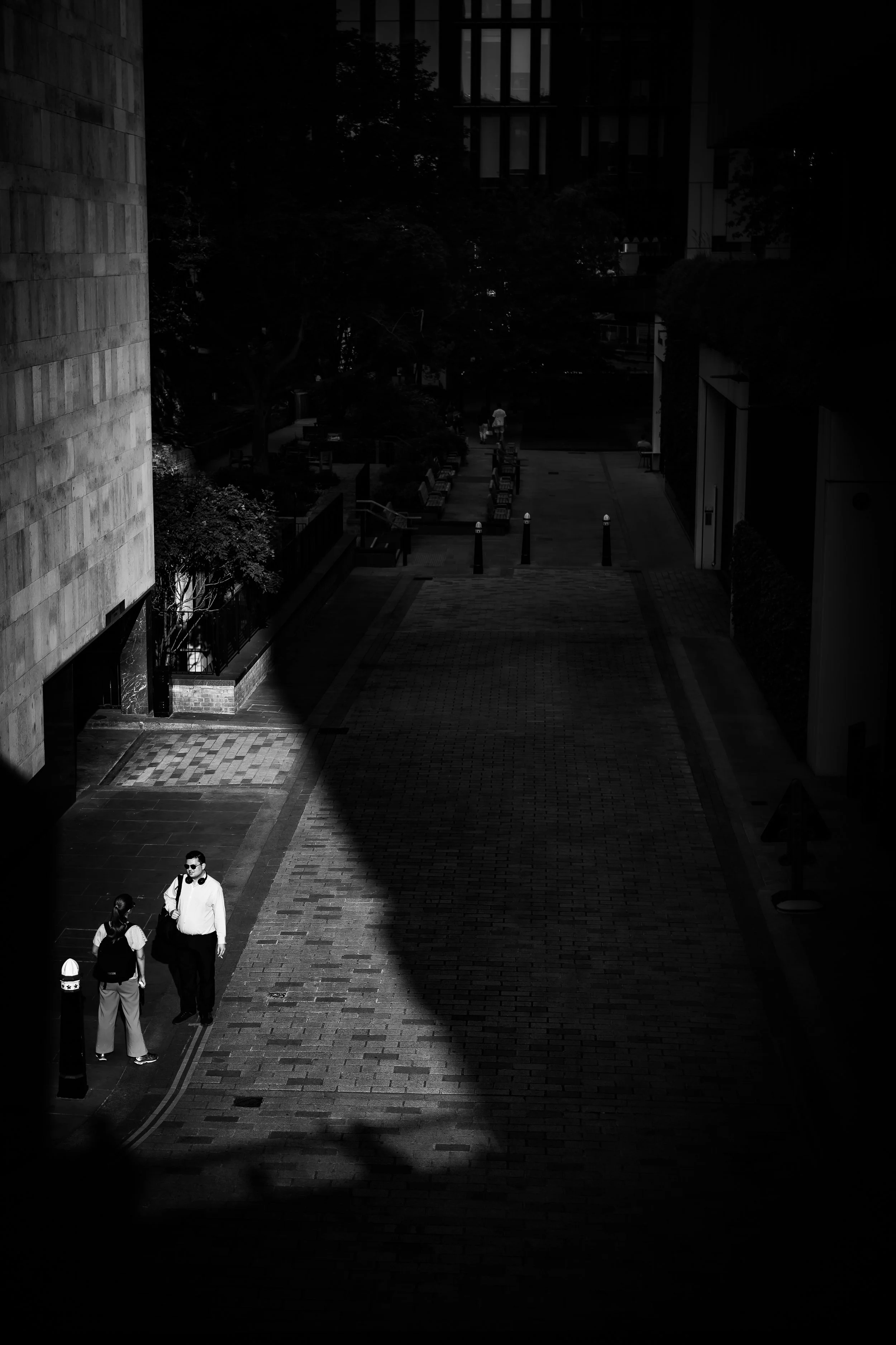 A black and white photo of an urban street scene with two pedestrians, a man and a woman, walking on the sidewalk. The sunlight creates a stark contrast, illuminating part of the sidewalk and the people while casting shadows.