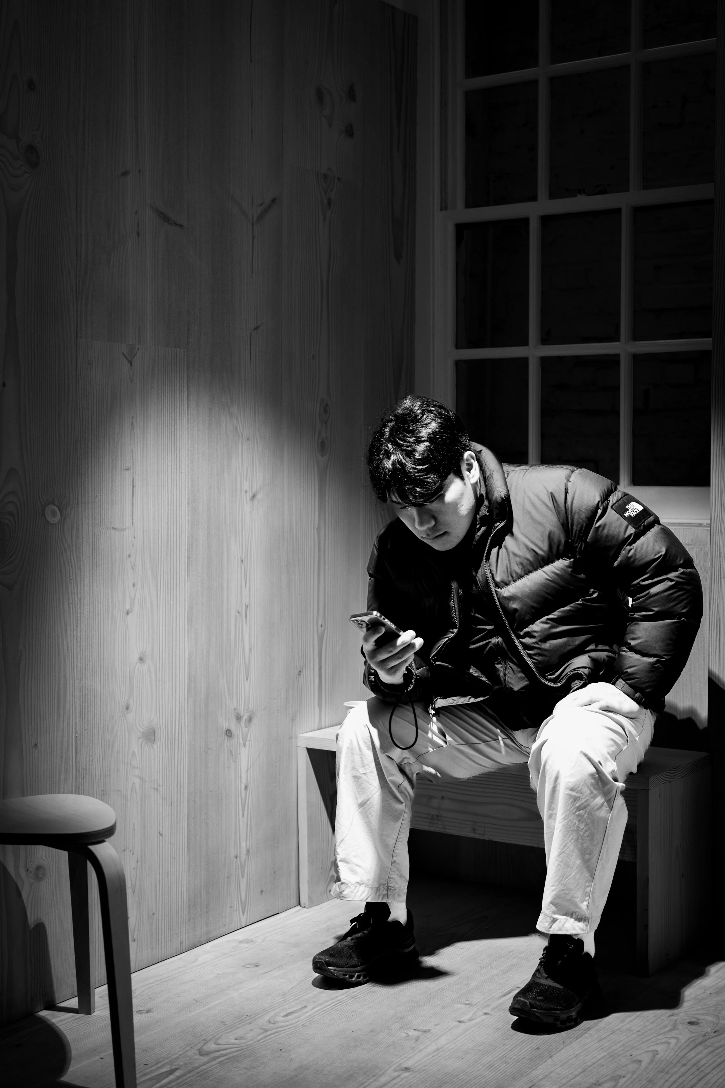 A young man wearing a North Face jacket and khaki pants is sitting on a wooden bench in a room with wood-paneled walls. He is looking at his smartphone and wearing hiking boots. There is a small stool and a chair beside him.