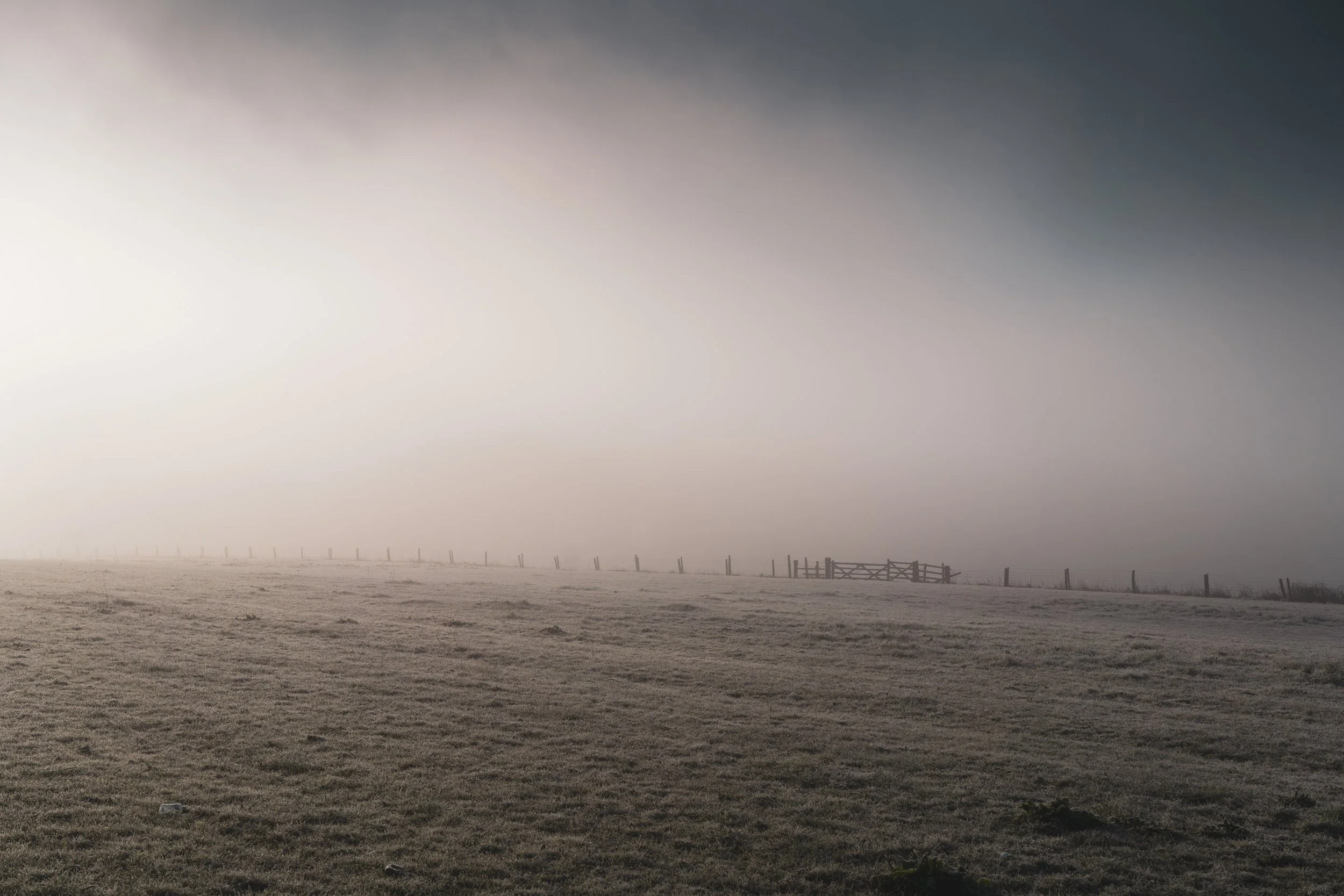 A foggy rural landscape with a grassy field, a fence, and a wooden gate. The fog obscures the background and creates a hazy atmosphere.