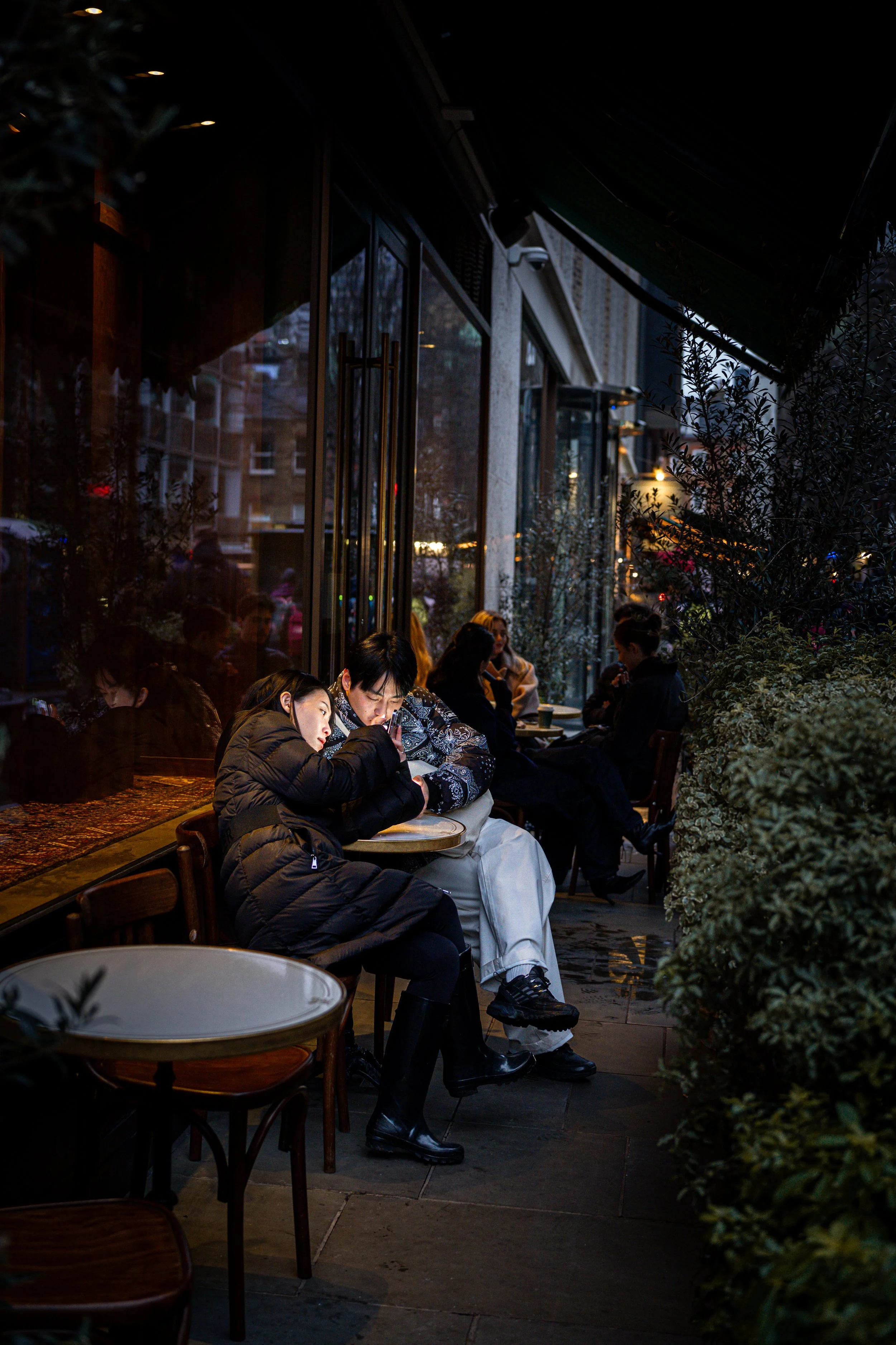 People sitting on outdoor patio at night, some looking at phones, surrounded by plants, with glass windows and building reflections visible.