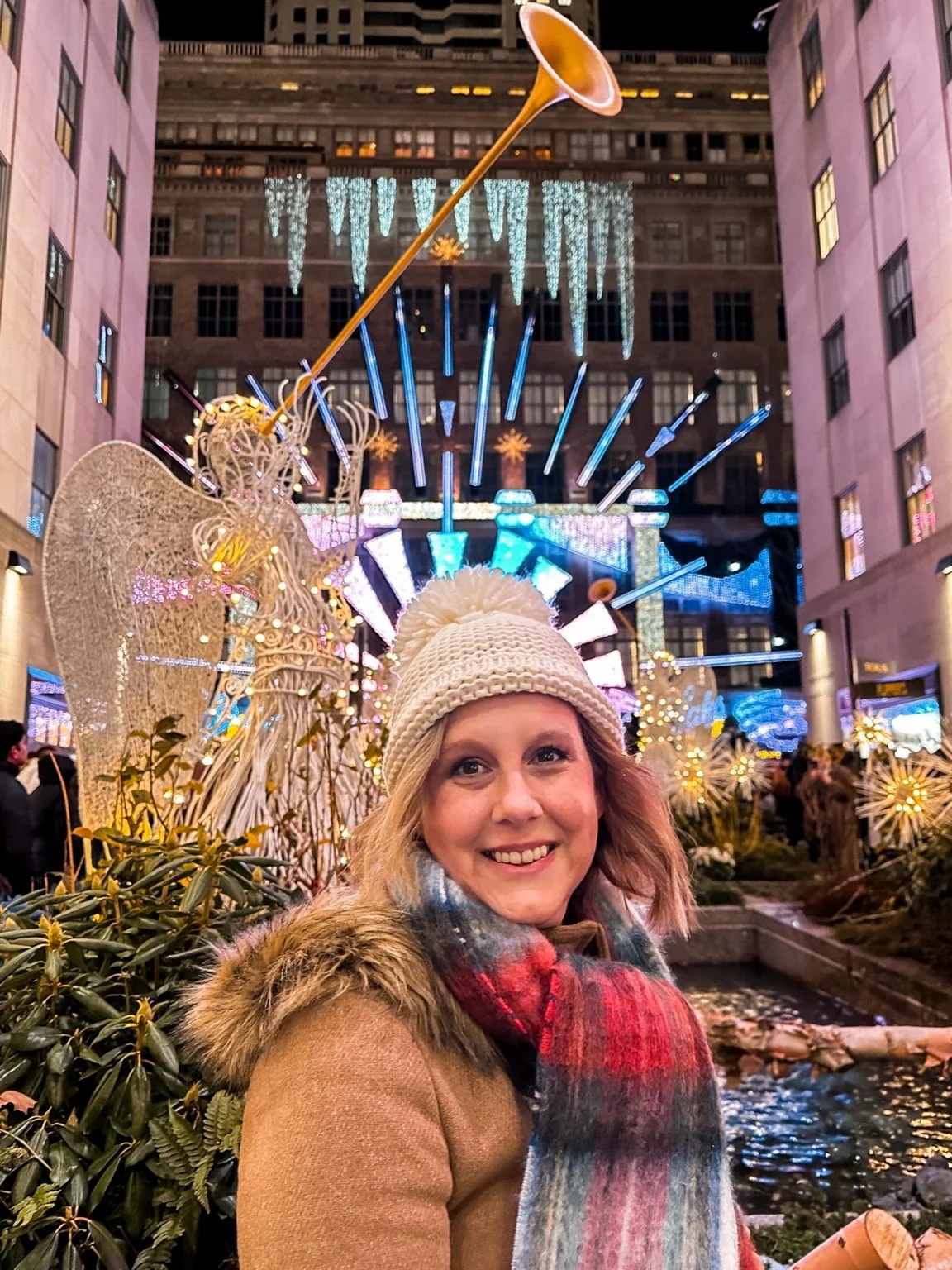 A smiling woman in a white knit hat and colorful scarf standing in front of holiday light displays and decorations in an urban setting at night.