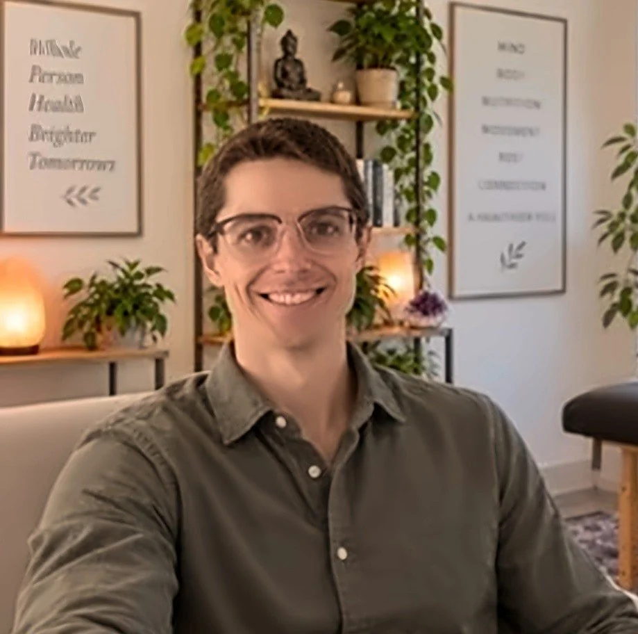 Dr. Mathew Richards, ND, naturopathic doctor at Transformative Medicine, smiling in his treatment room with plants and an acupuncture table behind him