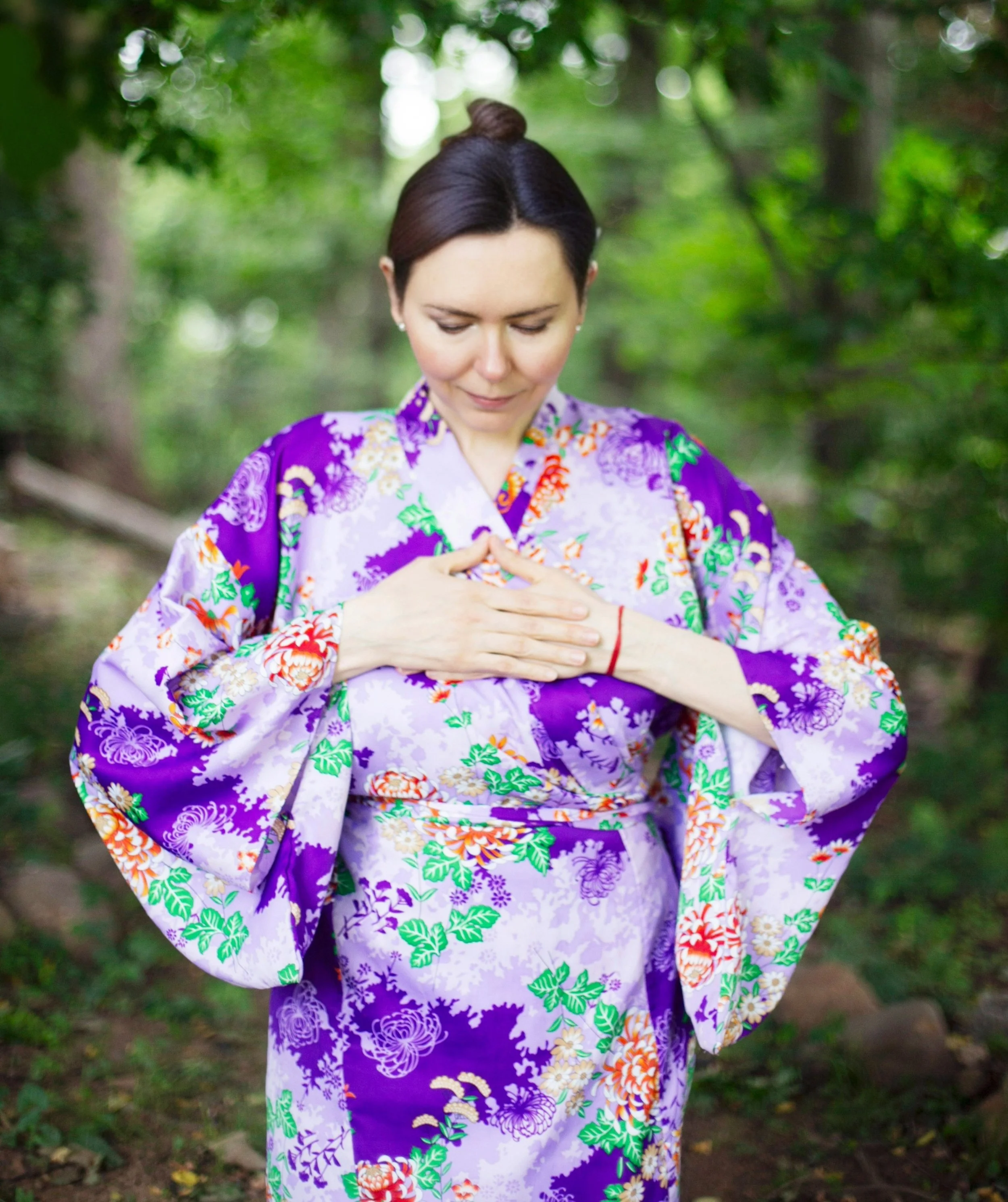 Dr. Lienna Wilson standing in a sunlit forest with eyes closed and hands resting over her heart, pausing for a moment of gratitude
