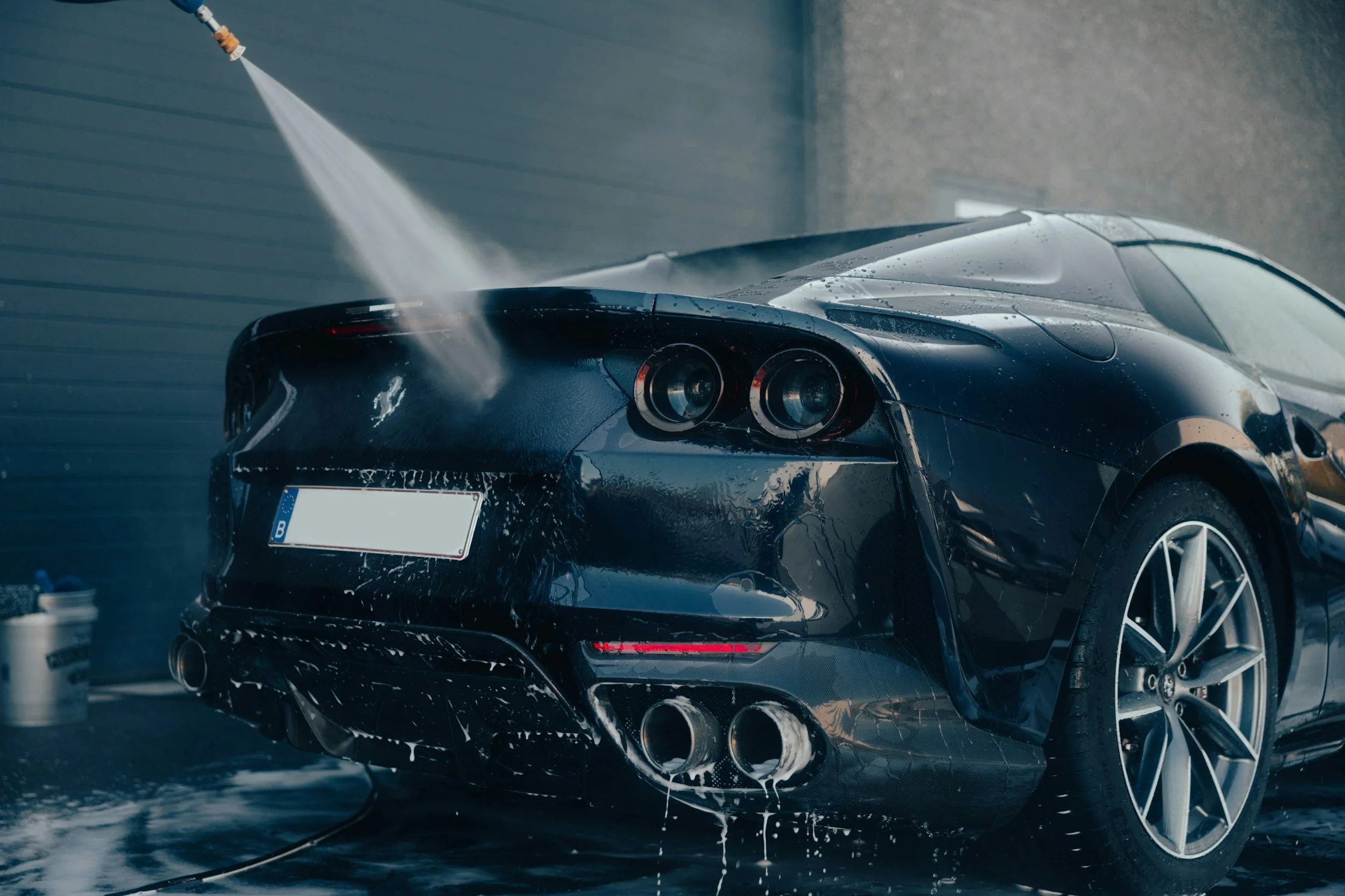 A black sports car is being washed with a foam spray on its rear end in a car wash bay. Water and soap suds are visible on the vehicle, with a hose nozzle aimed at the back. The car has a sleek design with visible tail lights and exhaust pipes.