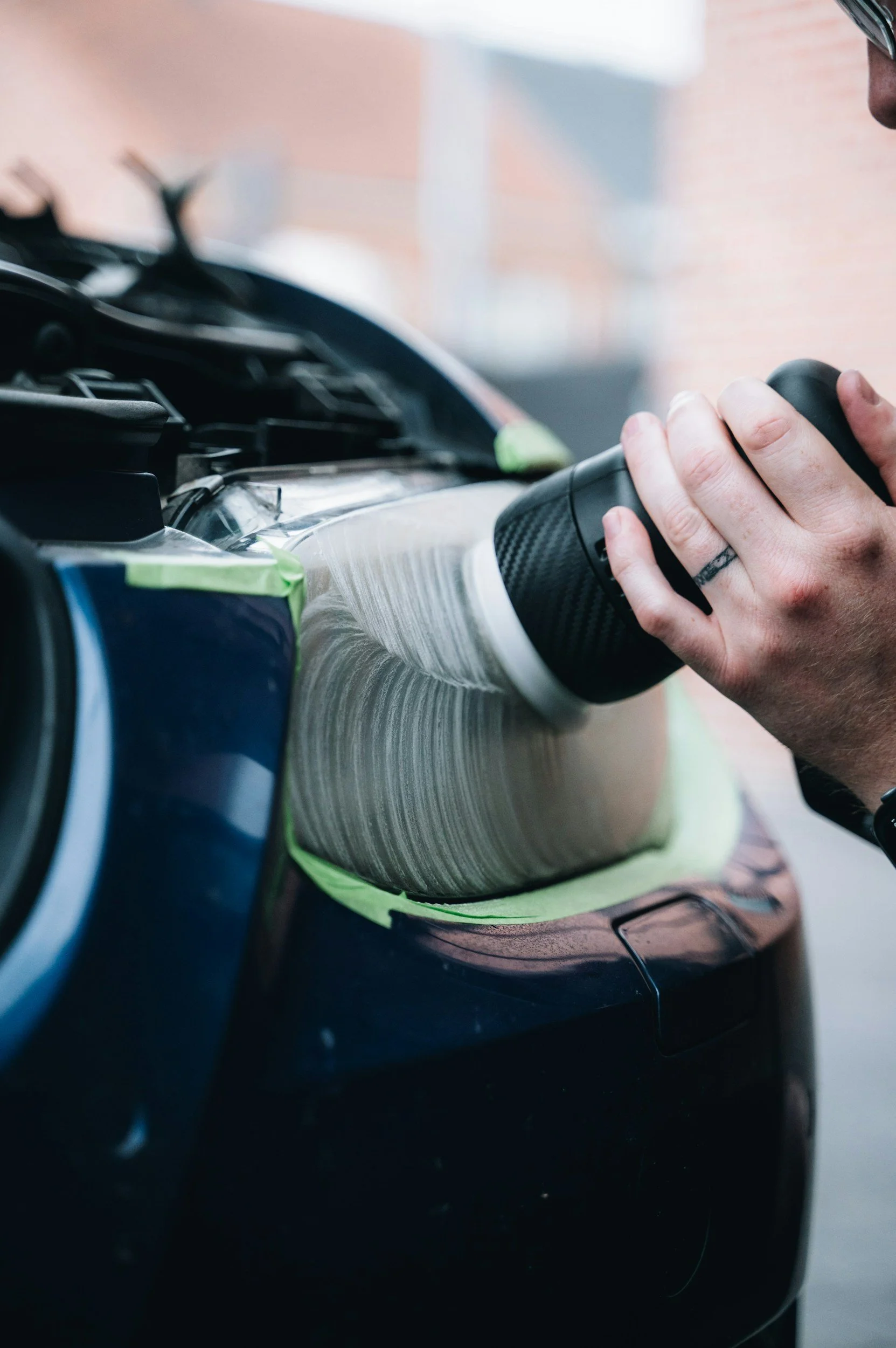 Person polishing a black car with a handheld polishing tool, wearing glasses, with a green protective cloth on the car.
