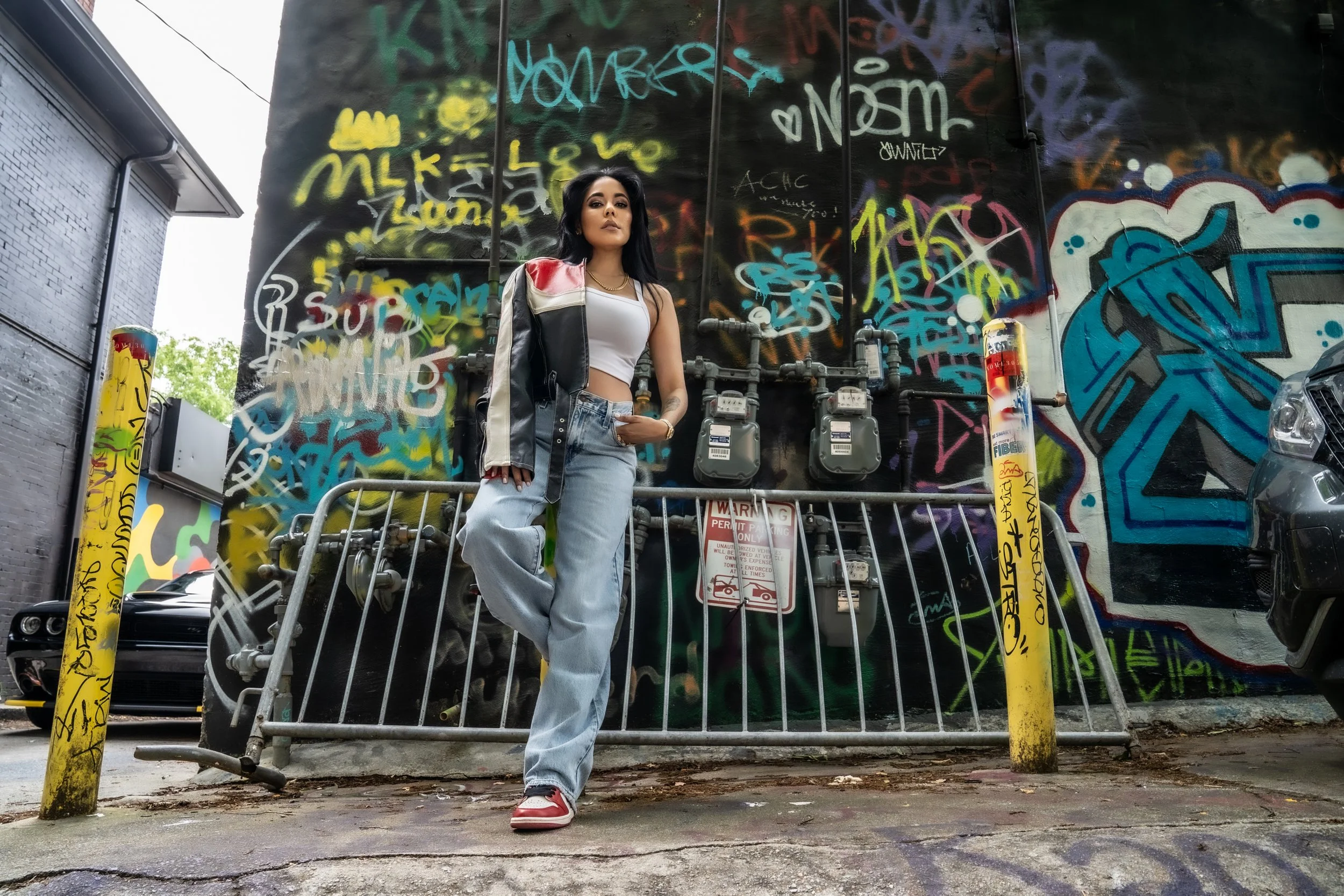 A woman with long dark hair wearing a white crop top, ripped jeans, and a black, red, and white leather jacket standing in front of a graffiti-covered wall. She poses with one hand on her hip and looks confidently forward. Graffiti and utility meters are visible behind her, with a parking lot and trees in the background.