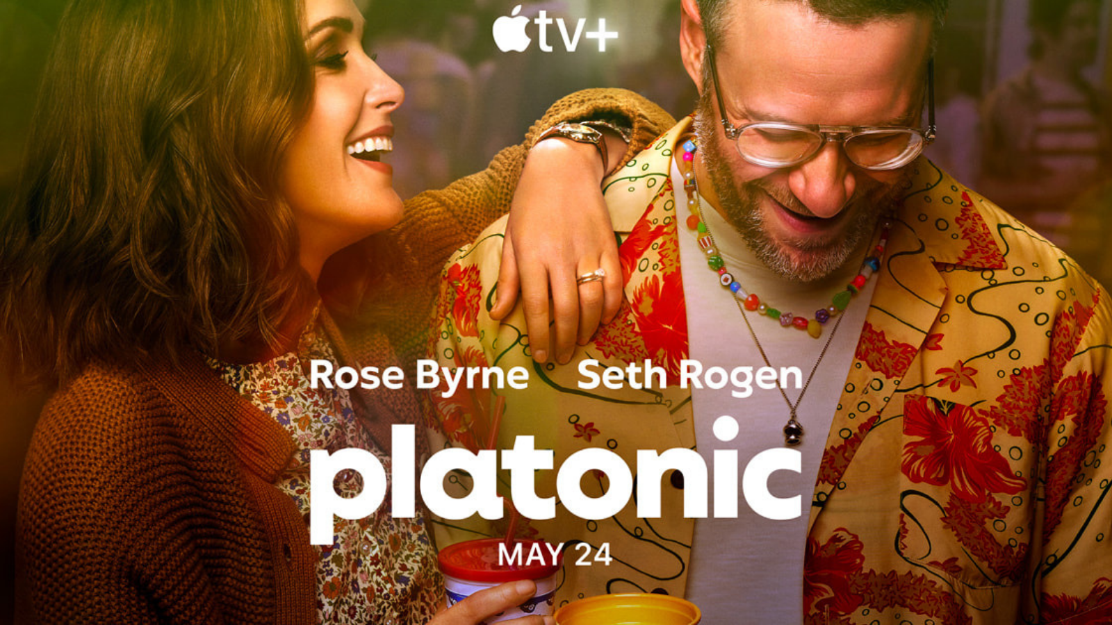 Two people smiling and enjoying each other's company at a colorful party or gathering, with text overlay promoting the movie 'Platonic' starring Rose Byrne and Seth Rogen, premiering on May 24 on Apple TV+.