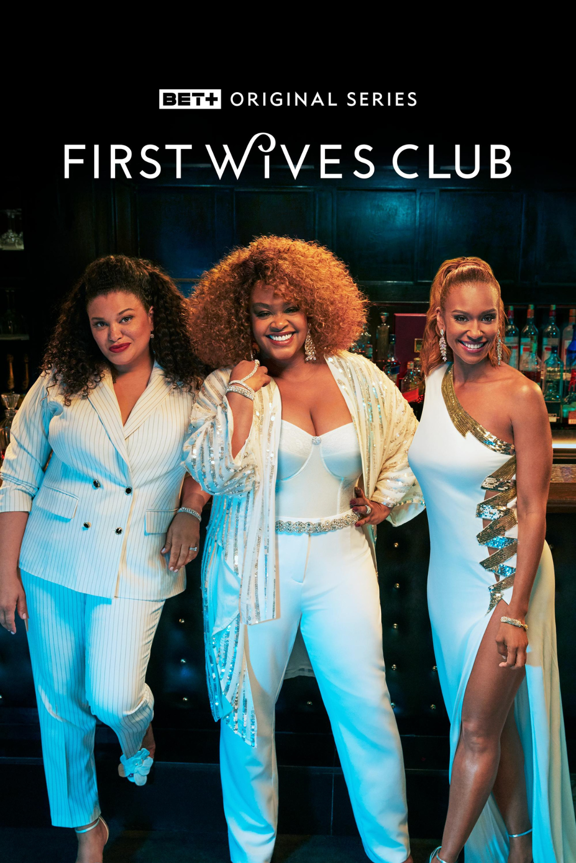 Three women in glamorous, stylish outfits posing together in a bar setting. Two women are wearing white outfits with gold accents, and the woman in the middle has curly hair and is dressed in white with metallic embellishments.