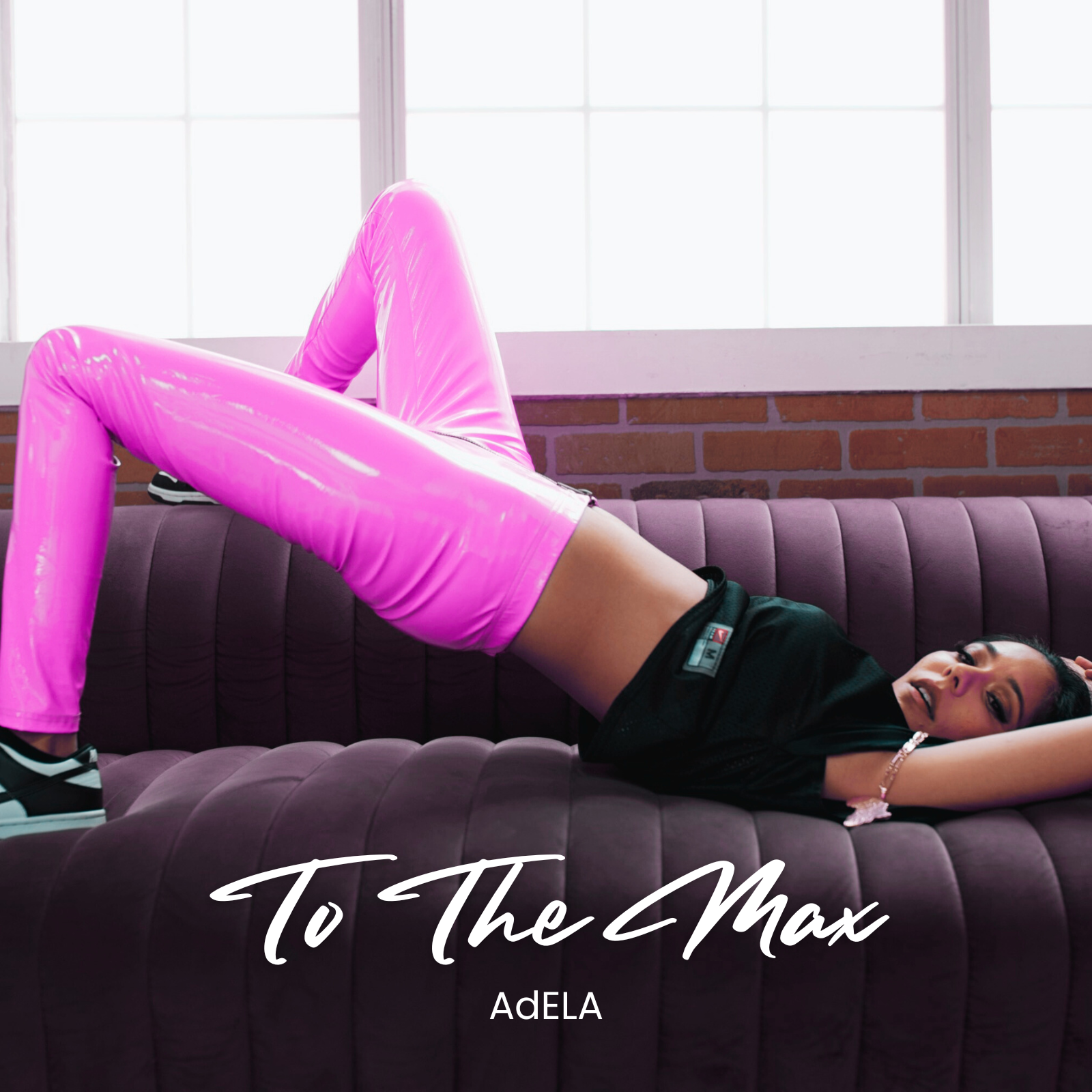 A woman lying on a purple velvet sofa, wearing shiny pink pants and black sneakers, with one arm behind her head in a relaxed pose. The background shows a large window with white frames and a brick wall.