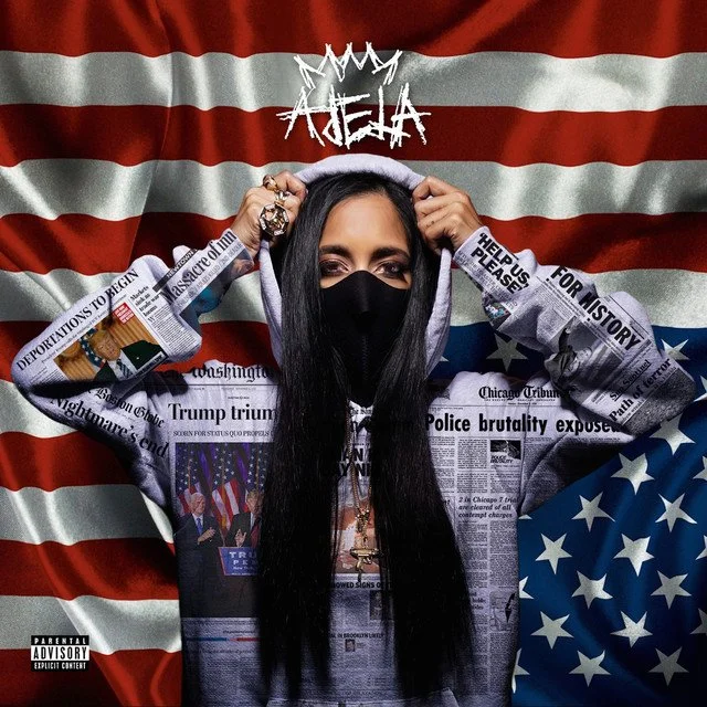 A woman wearing a black face mask and hoodie with a newspaper print design, holding her hood with both hands, standing in front of a large American flag with a dark red and white striped background and a blue field with white stars, with the word ATE