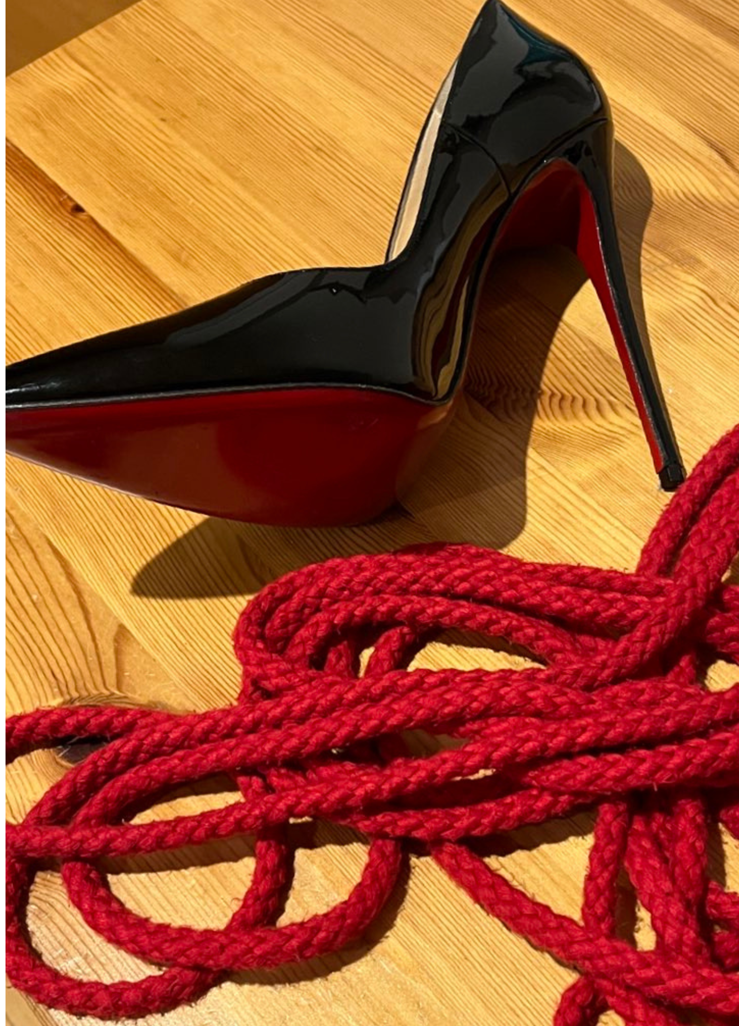 A Louboutin black high-heeled shoe with a red sole and a red rope on a wooden surface. Shibari.