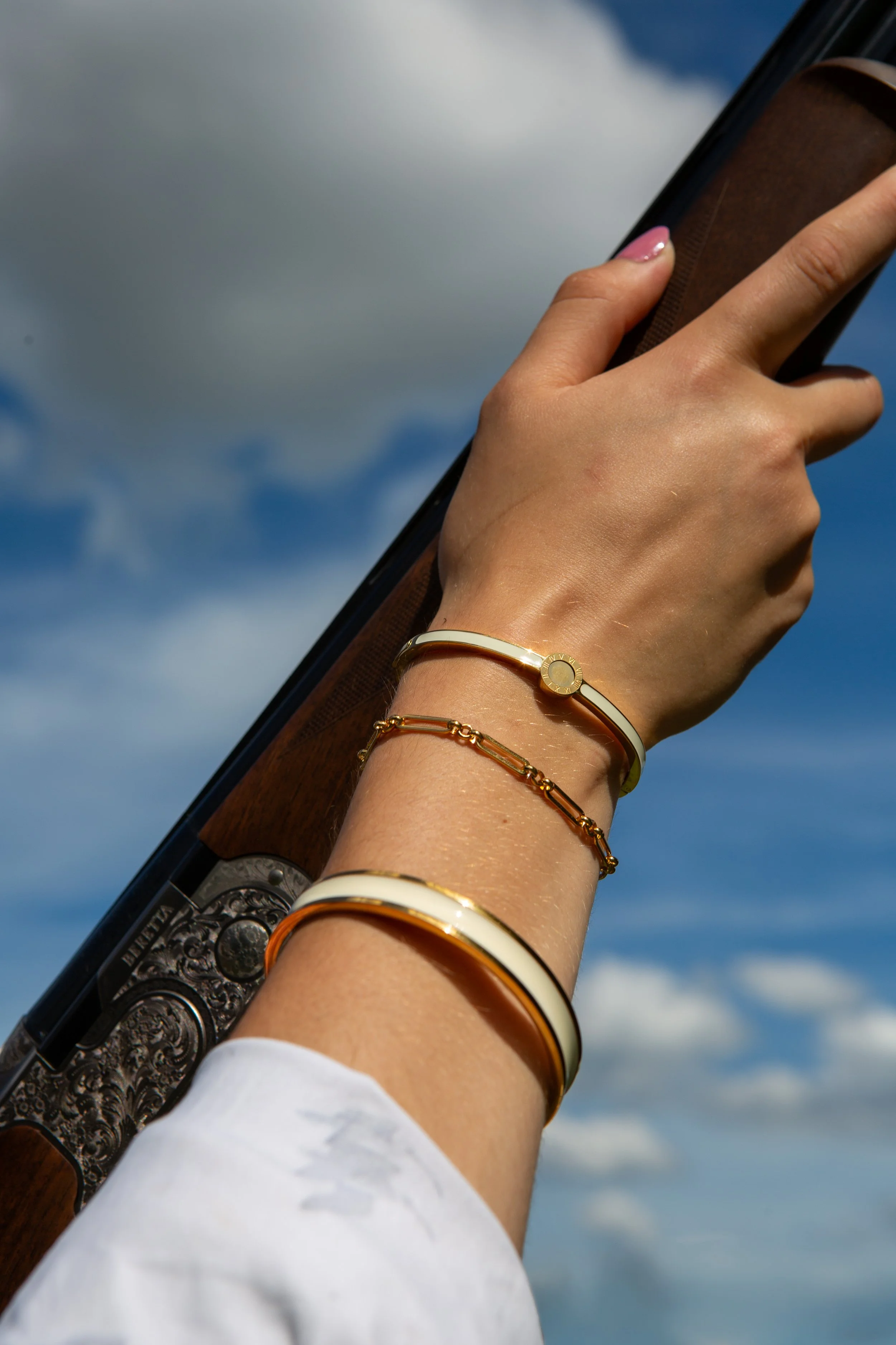 A person's hand holding a decorated pistol against a cloudy sky background, with jewelry including bracelets and a watch on the wrist.