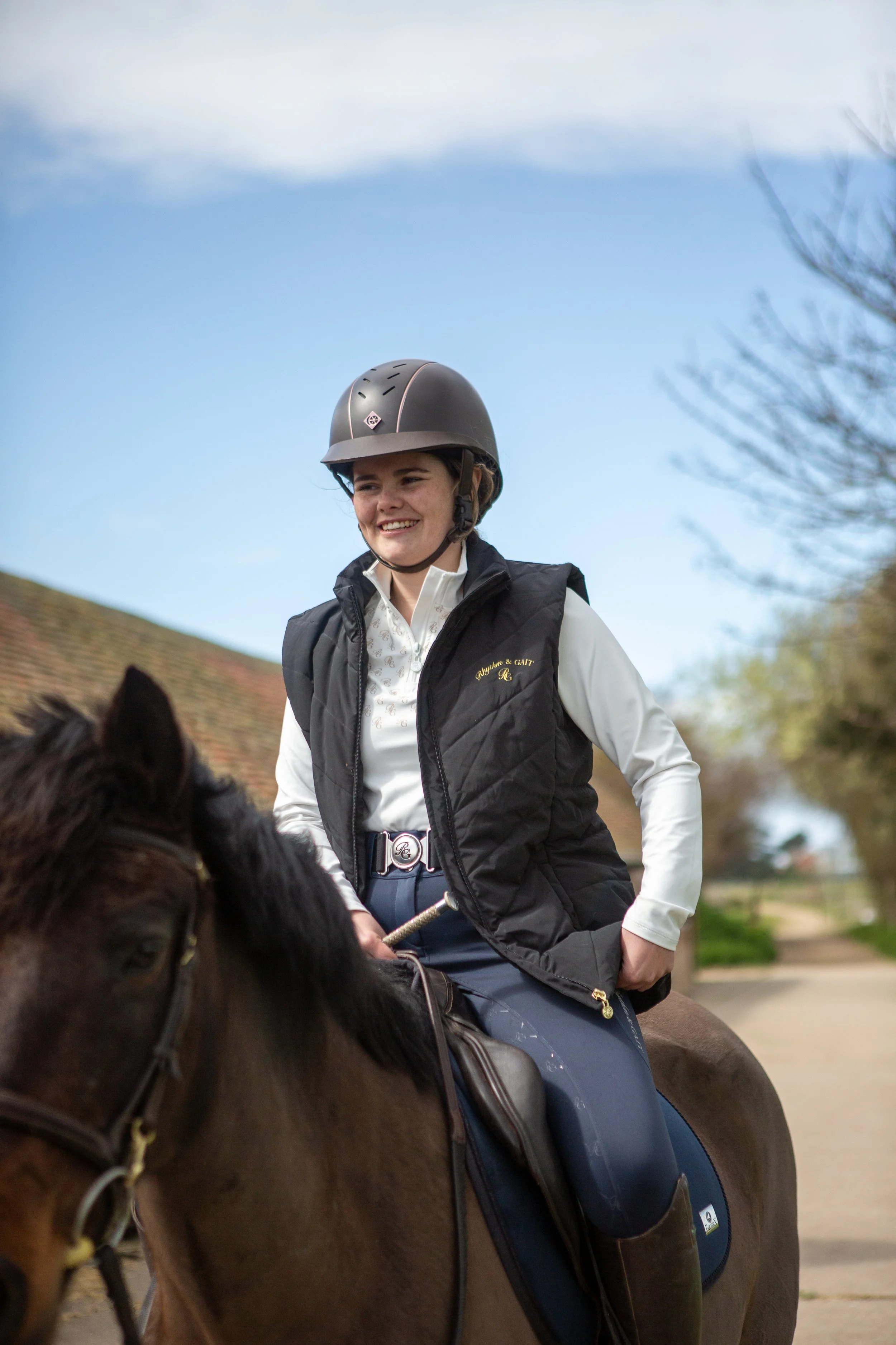 A young woman riding a horse outdoors, wearing a black riding helmet, a white collared shirt, and a black vest, smiling and holding the reins.