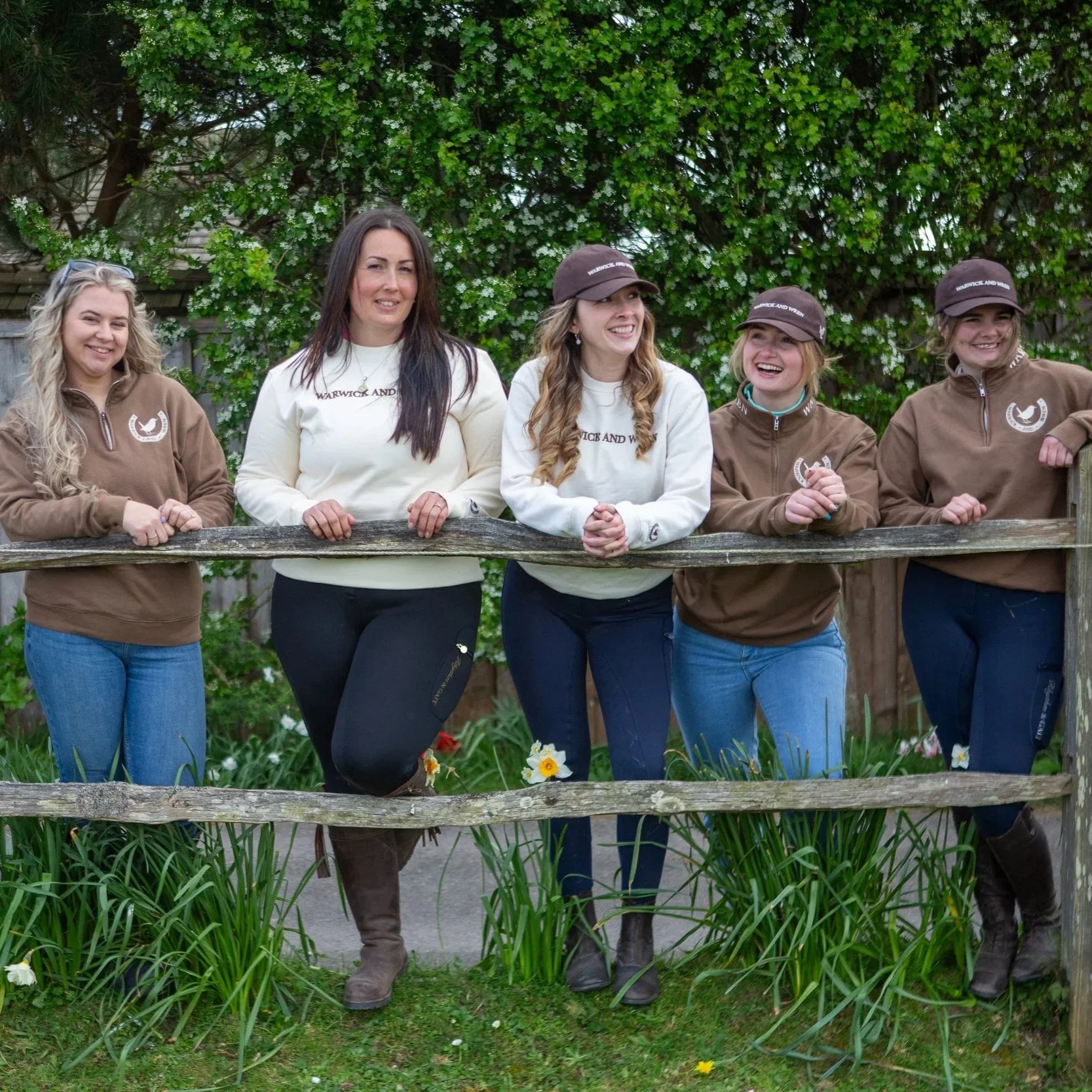Group of six women standing outdoors behind a wooden fence, smiling, wearing casual clothes and hats, with green trees and bushes in the background.