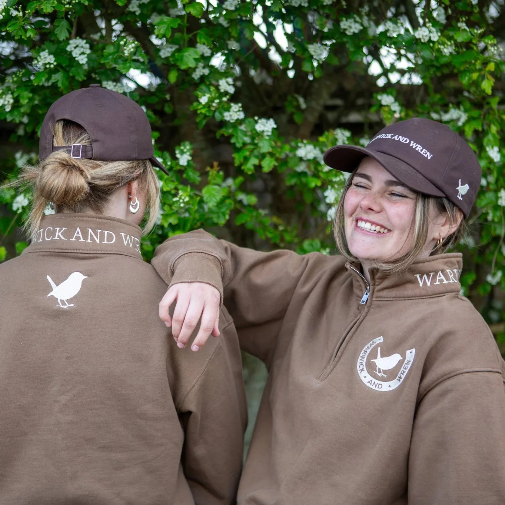 Two women wearing brown uniforms and caps with the logo of 'WICK AND WREN' embrace outdoors near a flowering bush with white blossoms, smiling and sharing a moment of camaraderie.