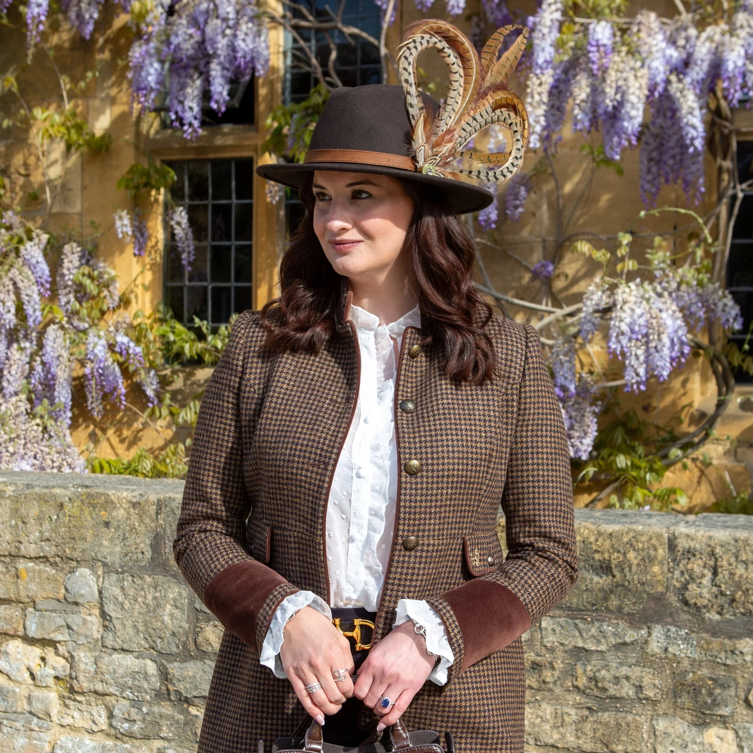 A woman standing outdoors in front of a stone wall covered with purple wisteria flowers, wearing a brown checkered coat, a white blouse, a wide-brimmed hat adorned with feathers, and holding a brown leather handbag.