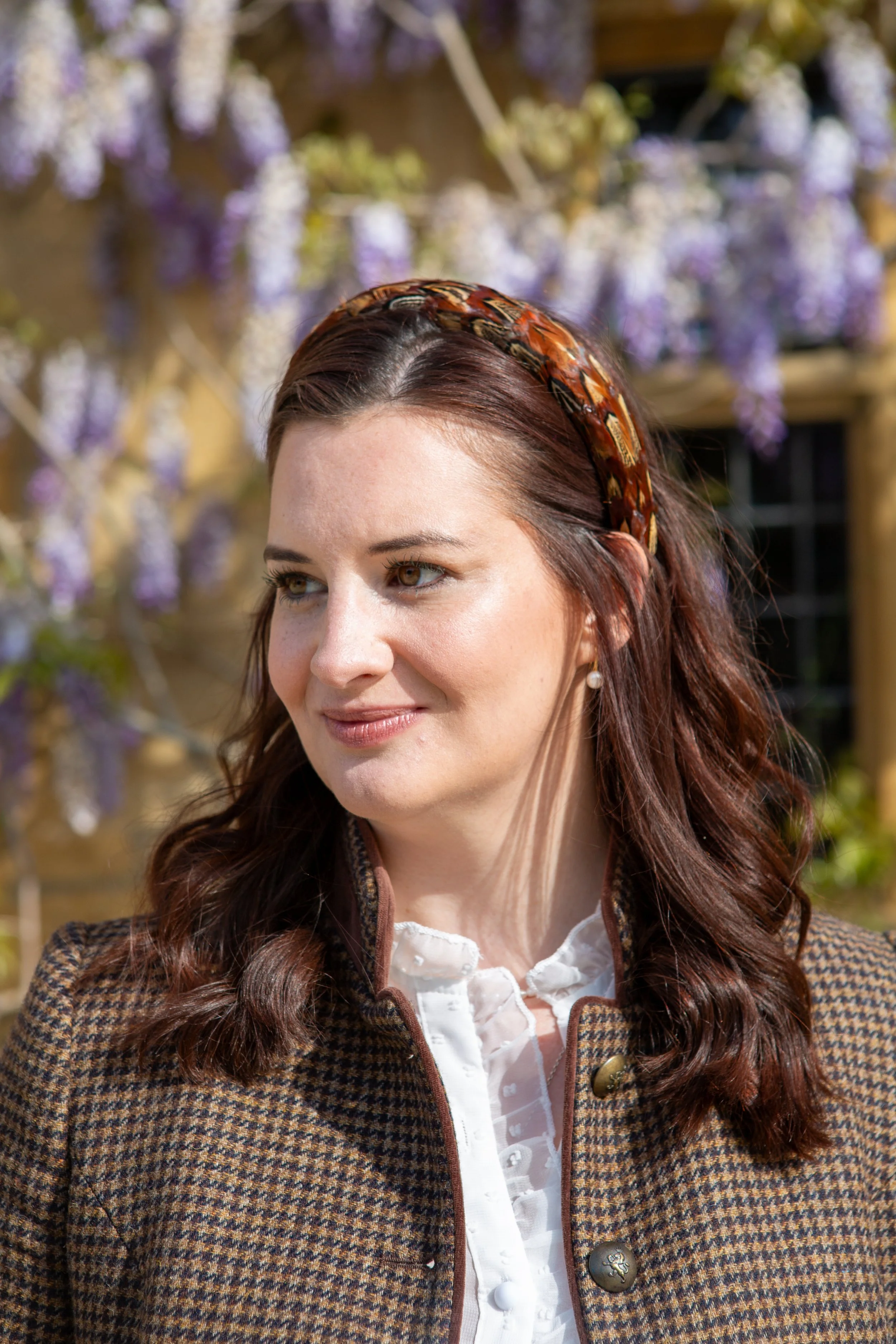 A woman with auburn hair and a headband, wearing a houndstooth blazer and pearl earrings, smiling outdoors with purple flowers in the background.