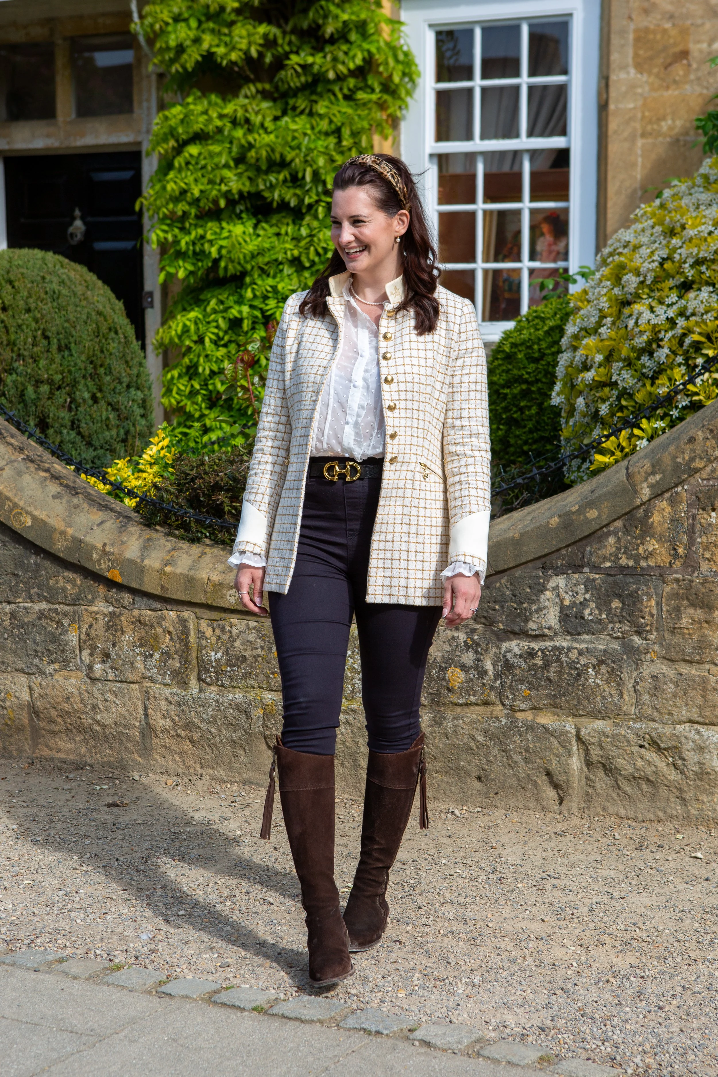 Woman smiling outdoors in front of a garden wall, wearing a cream blazer with gold buttons, a white blouse, black pants, and tall brown boots, with a leopard print headband and pearl earrings.