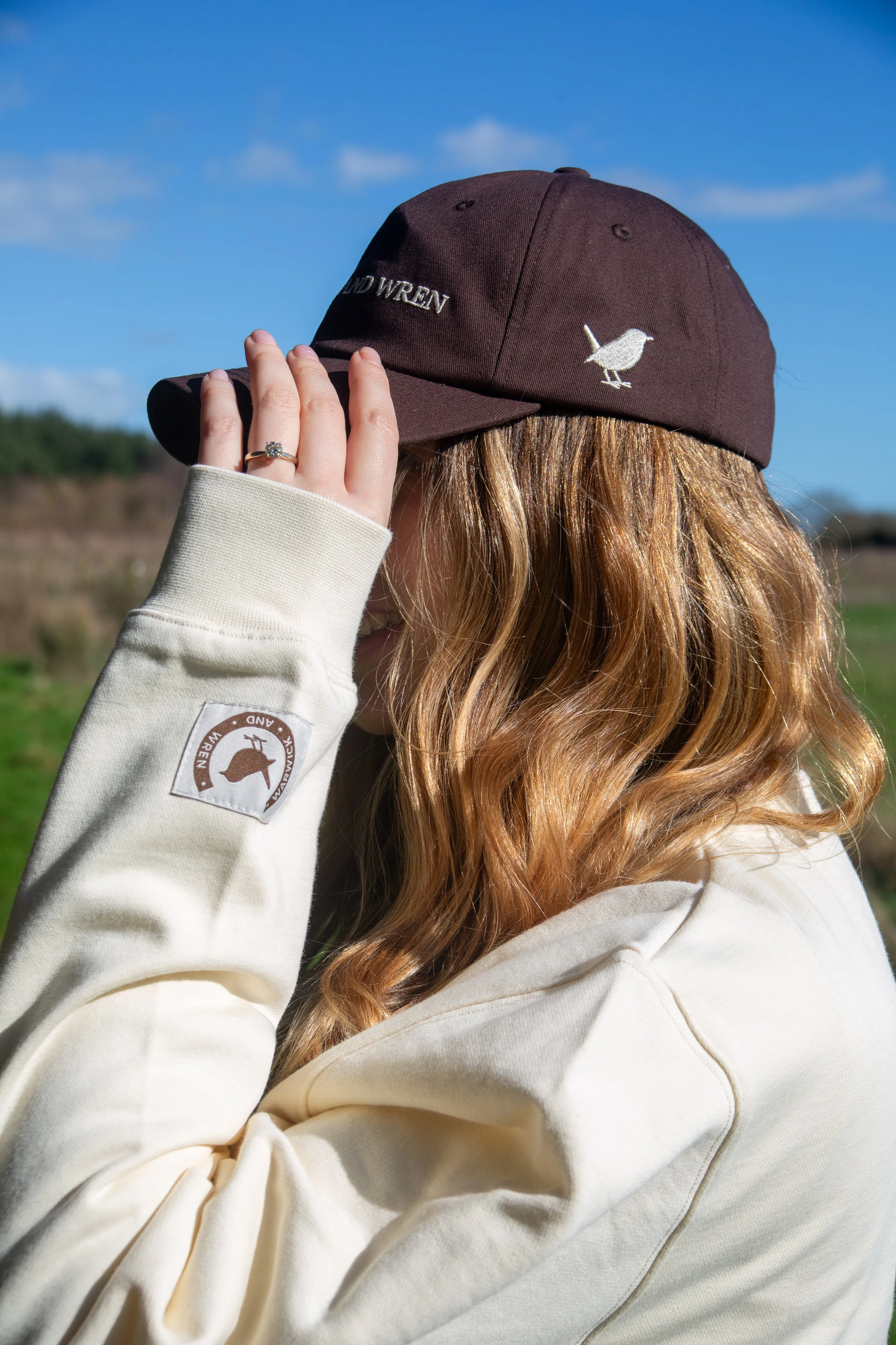 A person with long, wavy hair wearing a dark brown baseball cap with embroidered bird and text, a cream-colored hoodie with a logo patch on the sleeve, holding the brim of the cap with one hand, outdoors on a sunny day with a blue sky and some clouds