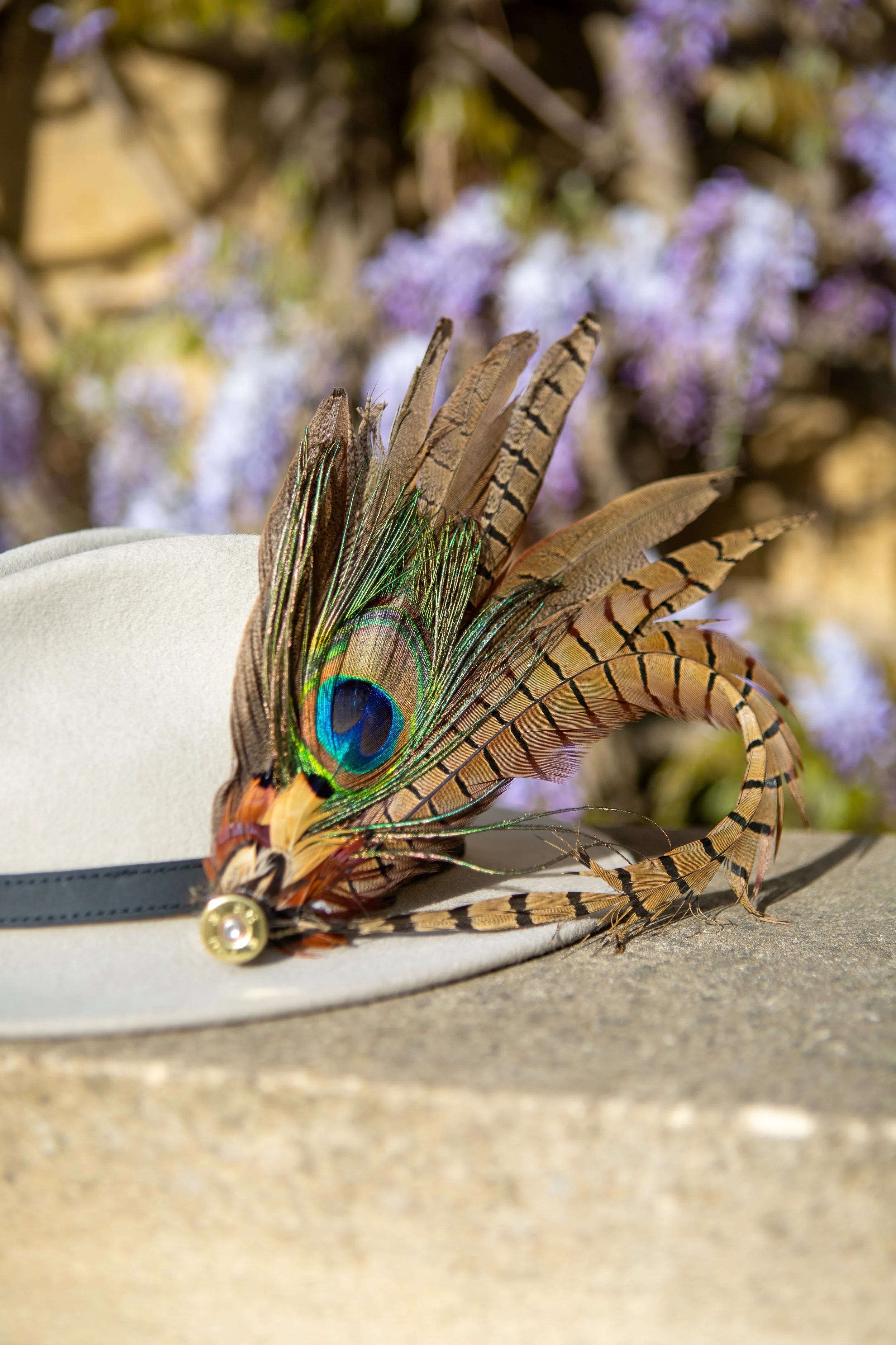 Decorative hat adorned with colorful peacock feathers and striped bird feathers, resting on a concrete surface with blurred purple flowers in the background.