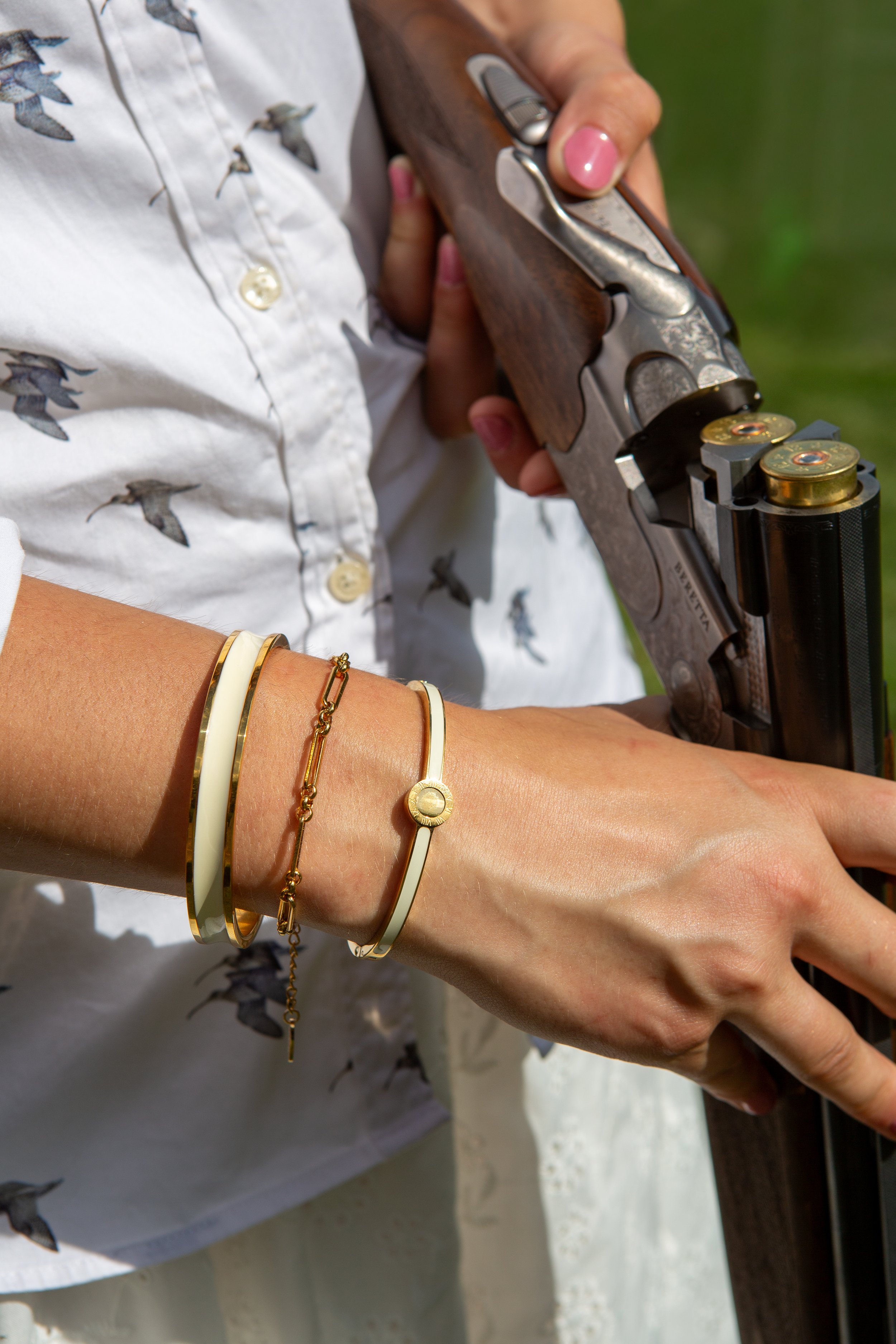 A person with light skin holding a revolver with a silver barrel and wooden handle. The person is wearing a white shirt with a bird pattern, a white bracelet, a gold bracelet, and a chain bracelet. The background is blurred green and outdoor setting.