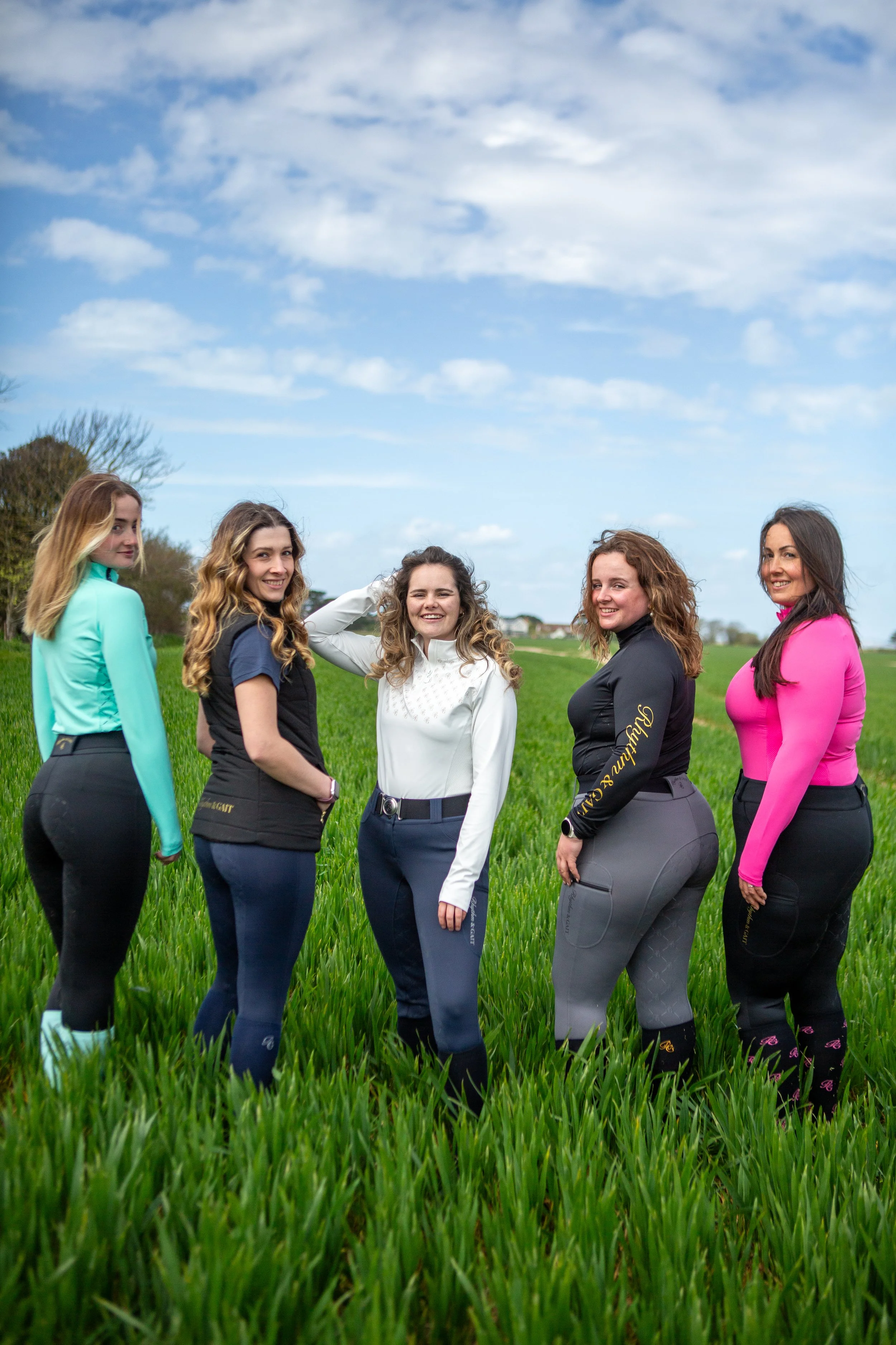 Six women standing in a grassy field under a partly cloudy sky, dressed in colorful athletic wear and smiling.