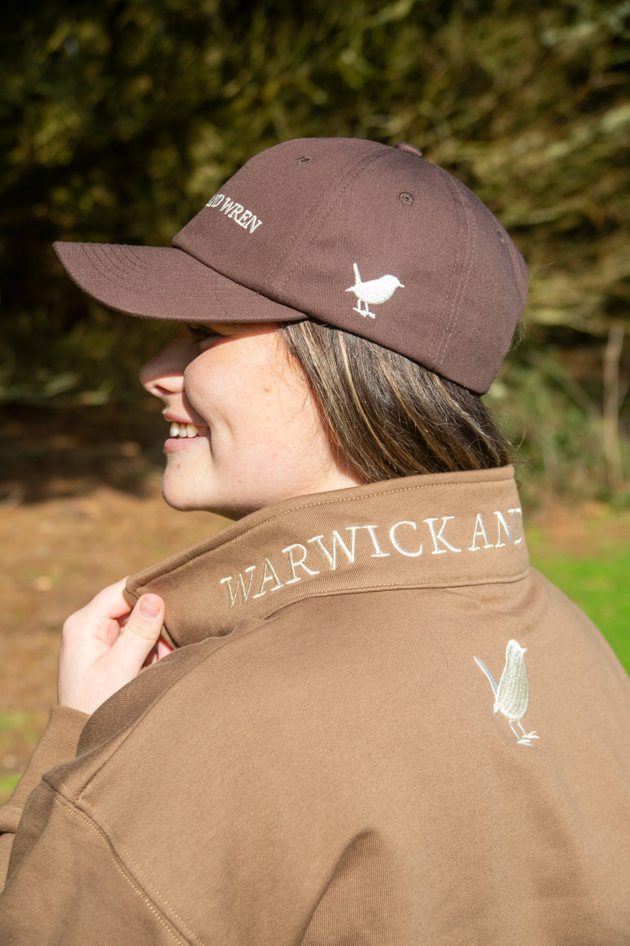 Side profile of a woman wearing a brown baseball cap with embroidered white bird and text, and a brown jacket with embroidered white text 'WARWICK AND'.