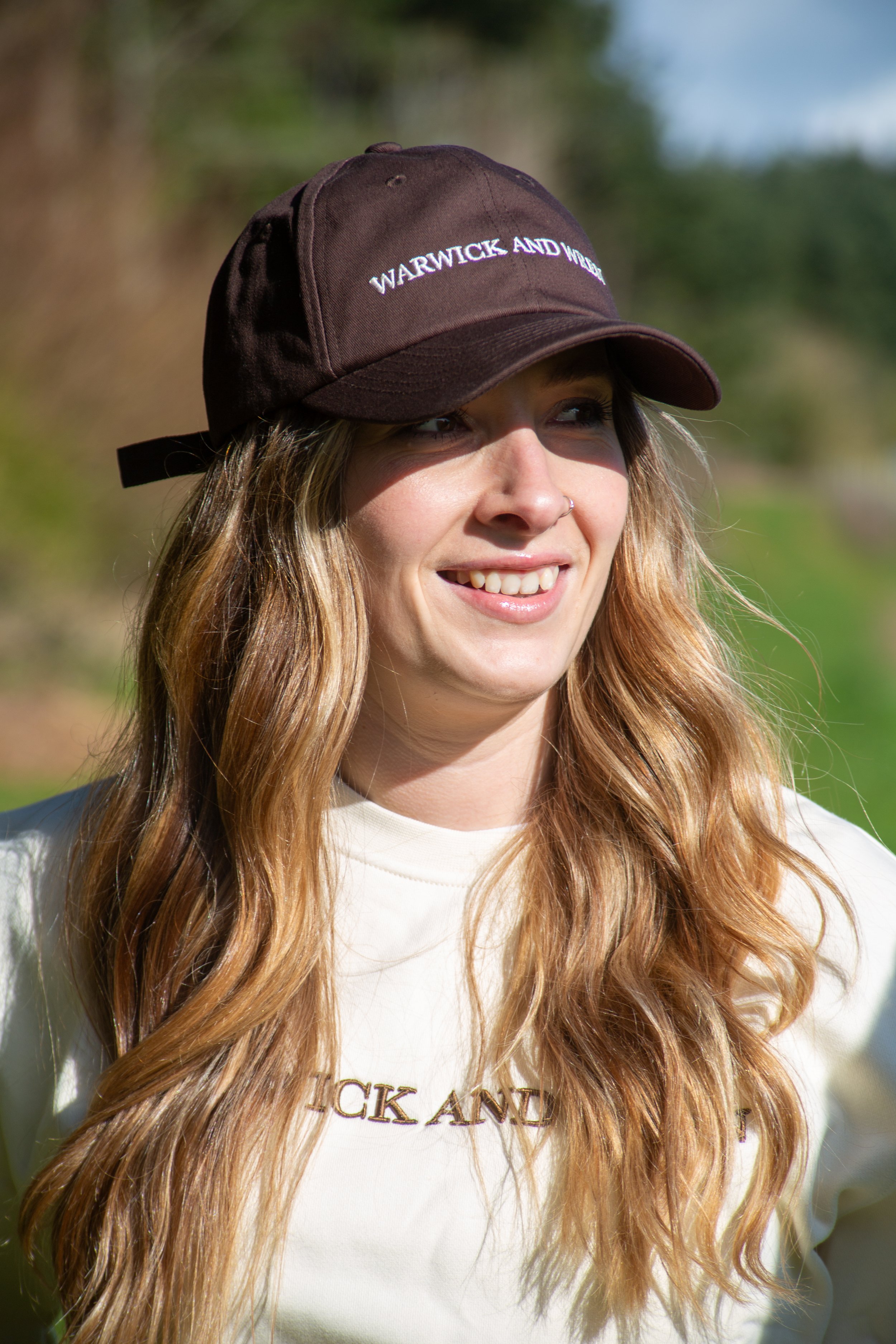 A young woman with long, wavy red hair wearing a brown baseball cap with the words 'WARWICK AND W' embroidered on it and a cream-colored T-shirt with text that is partially out of the frame, standing outdoors on sunny day with blurred greenery and a 