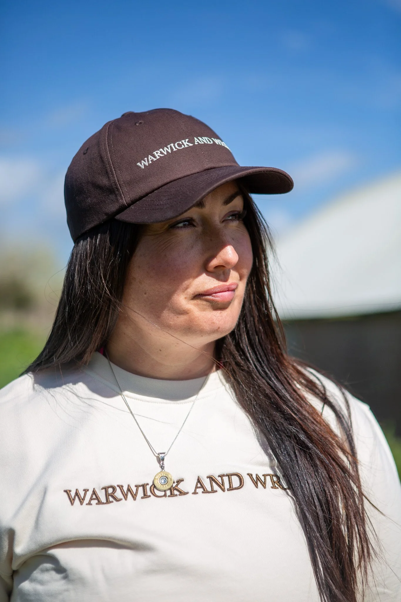 Close-up of a young woman with long dark hair wearing a brown Warwick and Warwick baseball cap, white shirt with logo, and a silver necklace, standing outdoors against a blue sky.