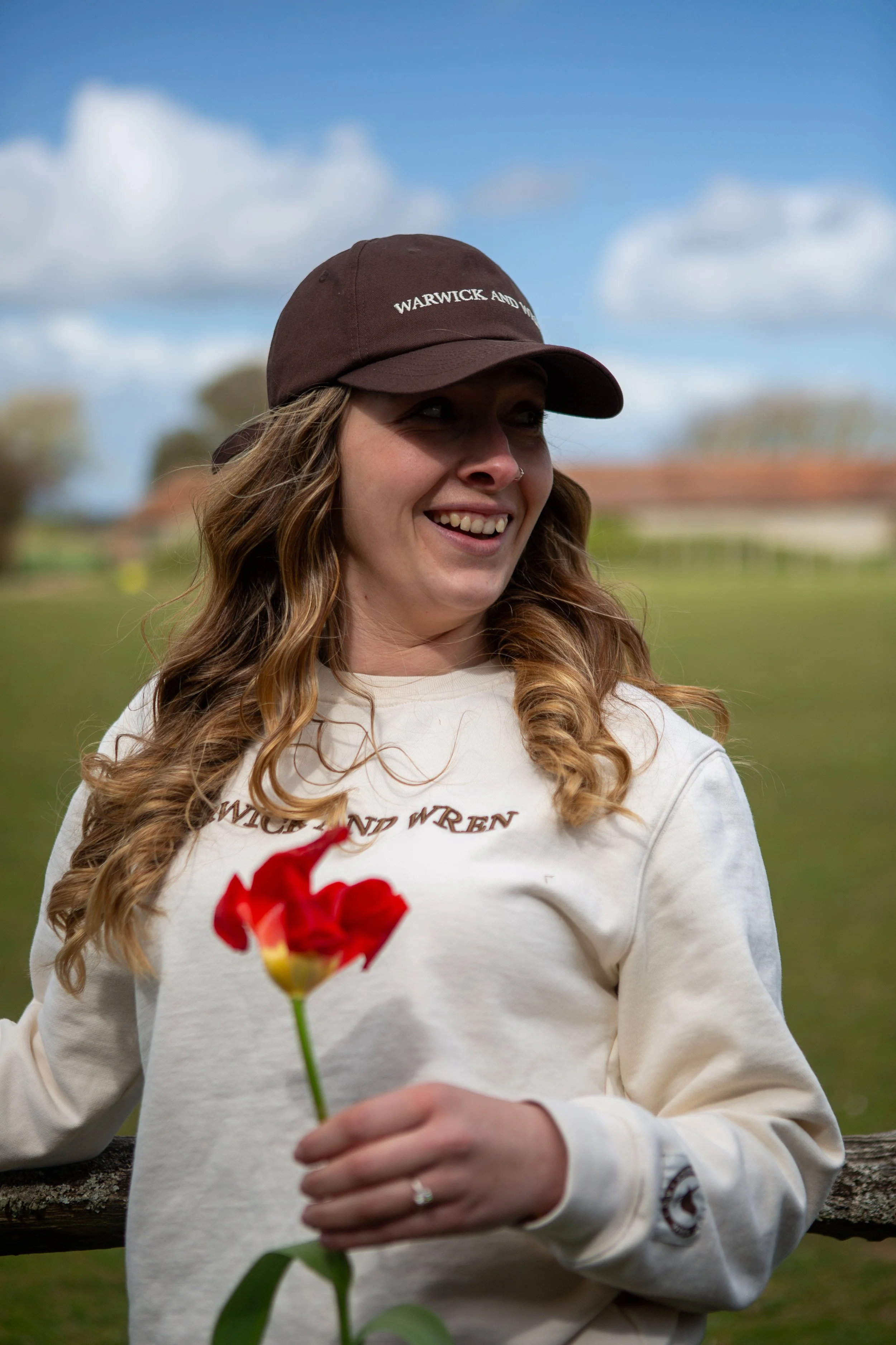 A woman with long curly hair wearing a brown cap and a cream-colored sweatshirt holds a red flower, standing outdoors with a cloudy sky and blurred trees in the background.
