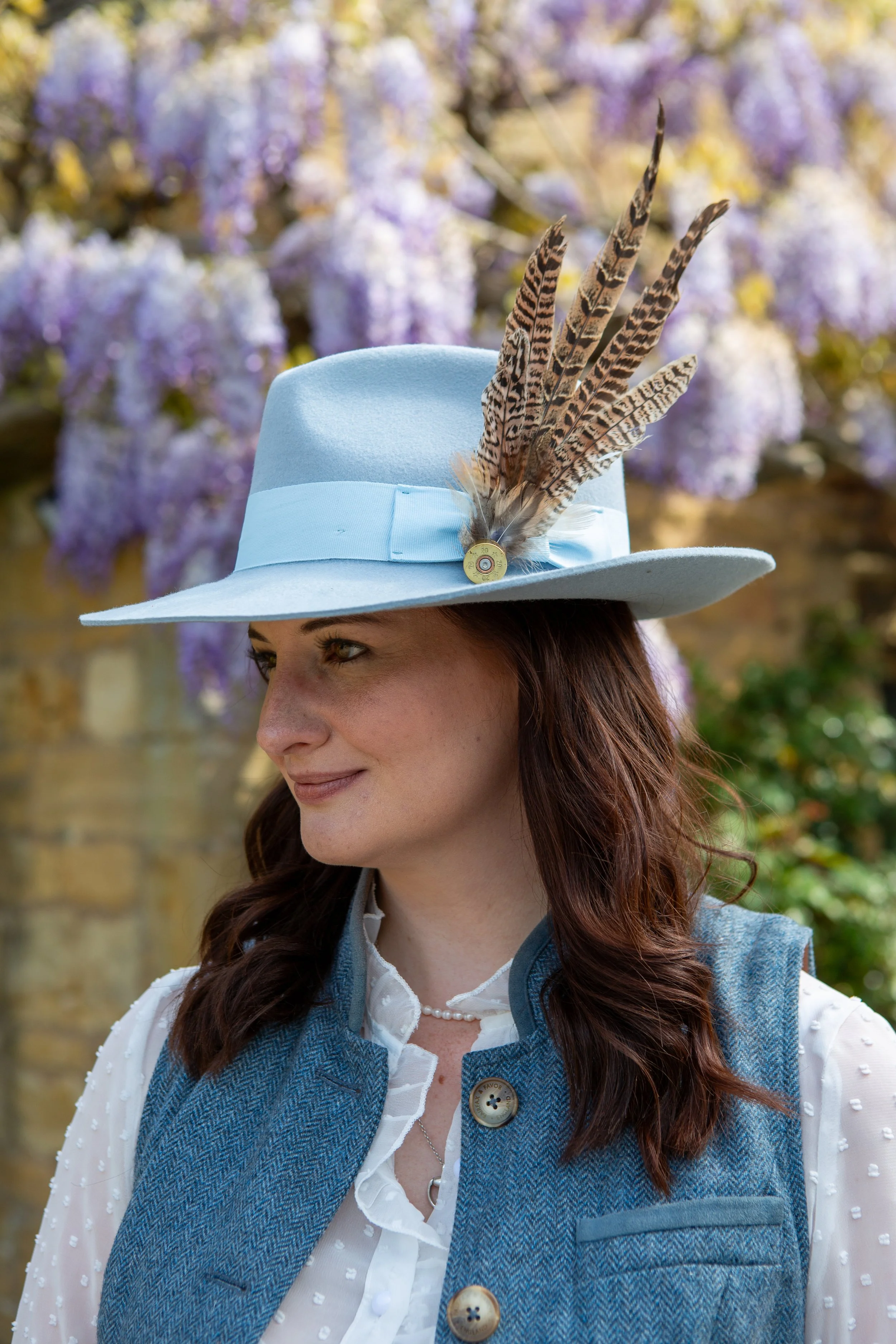 A woman wearing a light blue hat with feathers and a button, a white blouse, and a blue vest. She is outdoors with purple flowering trees in the background.