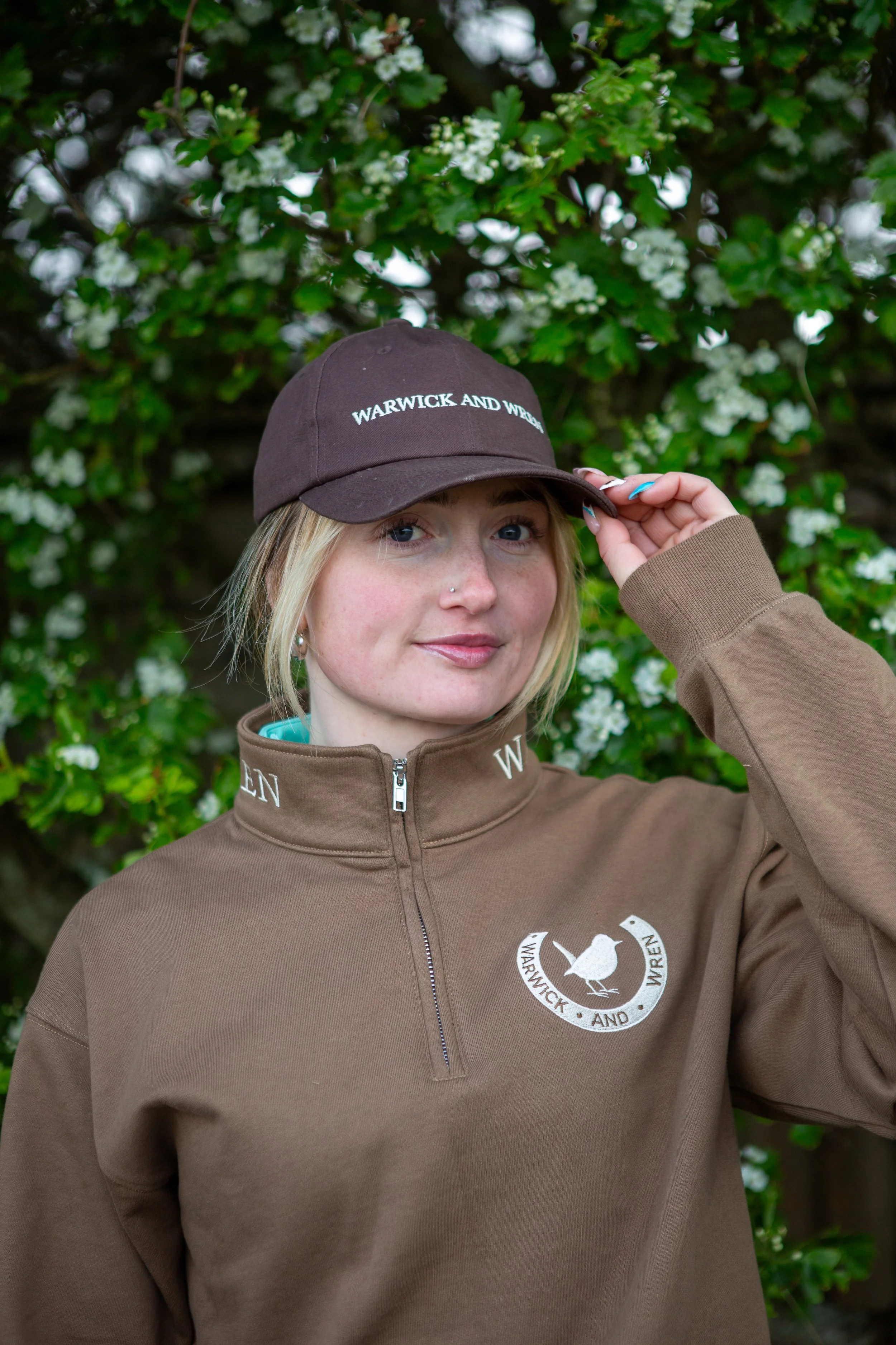 A young woman with blonde hair wearing a brown baseball cap, a brown jacket with a logo, and a nose piercing, standing in front of a bush with white flowers.