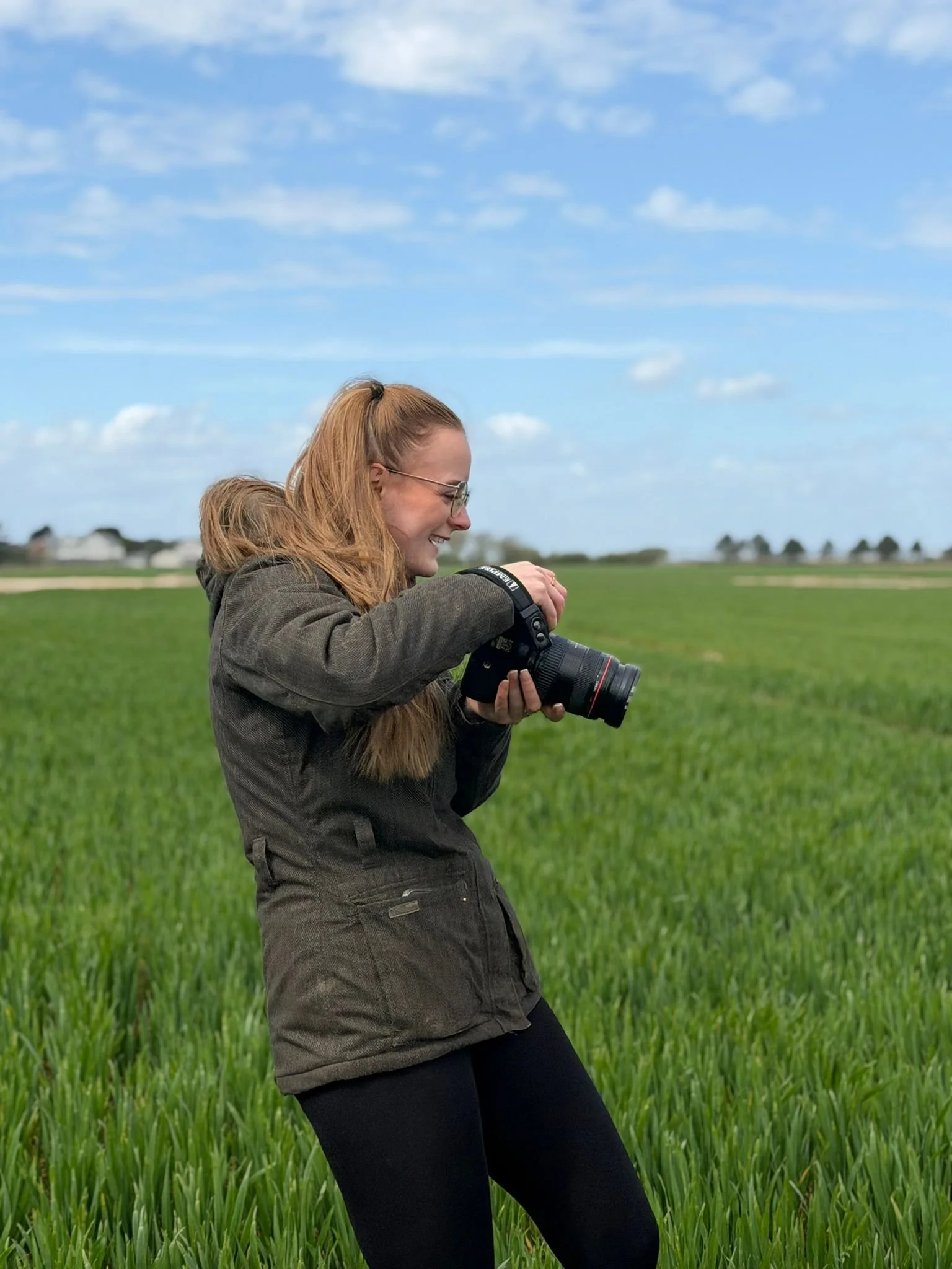 A woman with glasses and long reddish hair, wearing a brown jacket, stands in a green field while looking at her camera during daytime with a blue sky and a few clouds in the background.