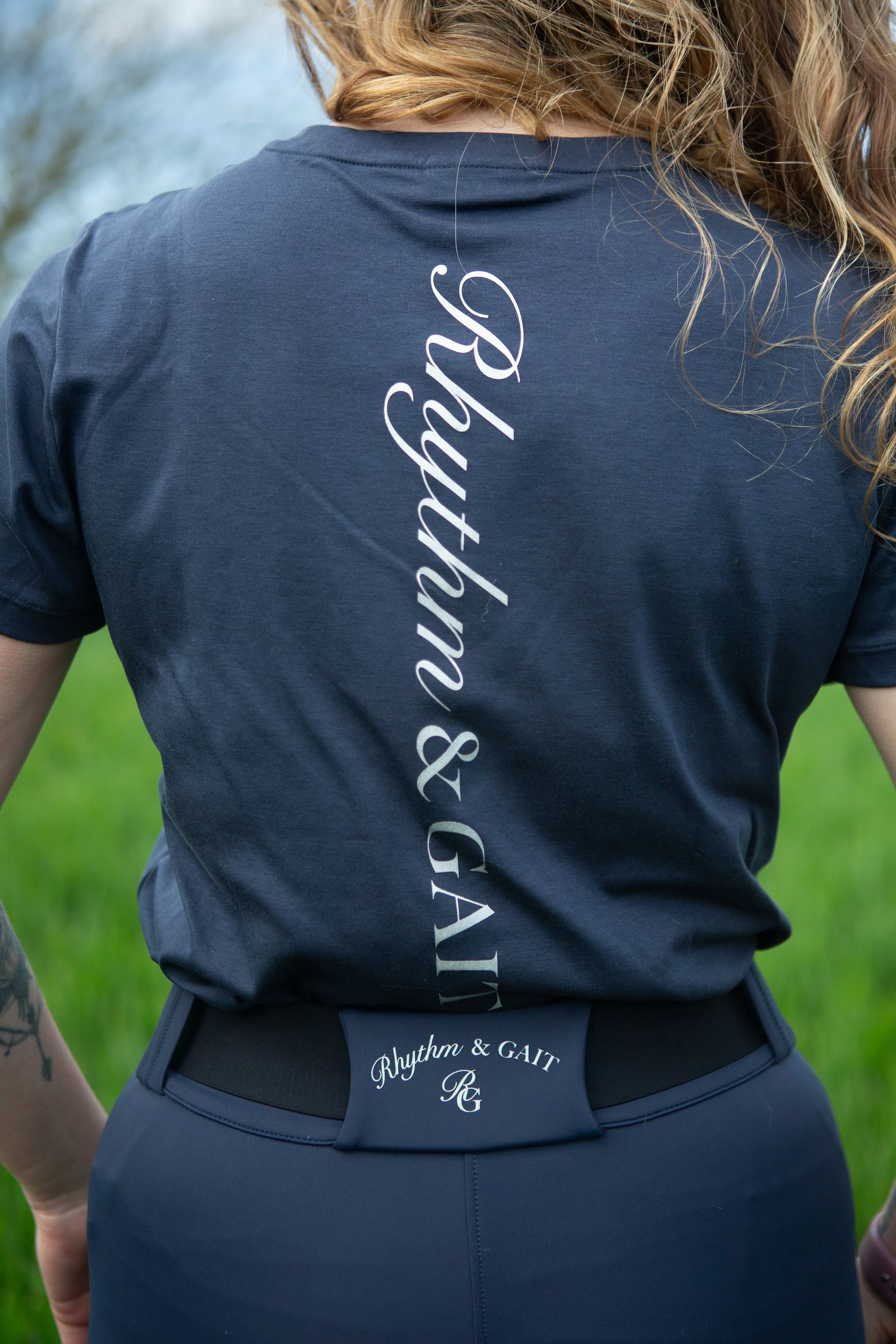 Close-up of a woman wearing a dark blue athletic t-shirt with white cursive writing and a black waist belt with the words "Rhythm & GAIT" embroidered on it, standing outdoors with greenery in the background.