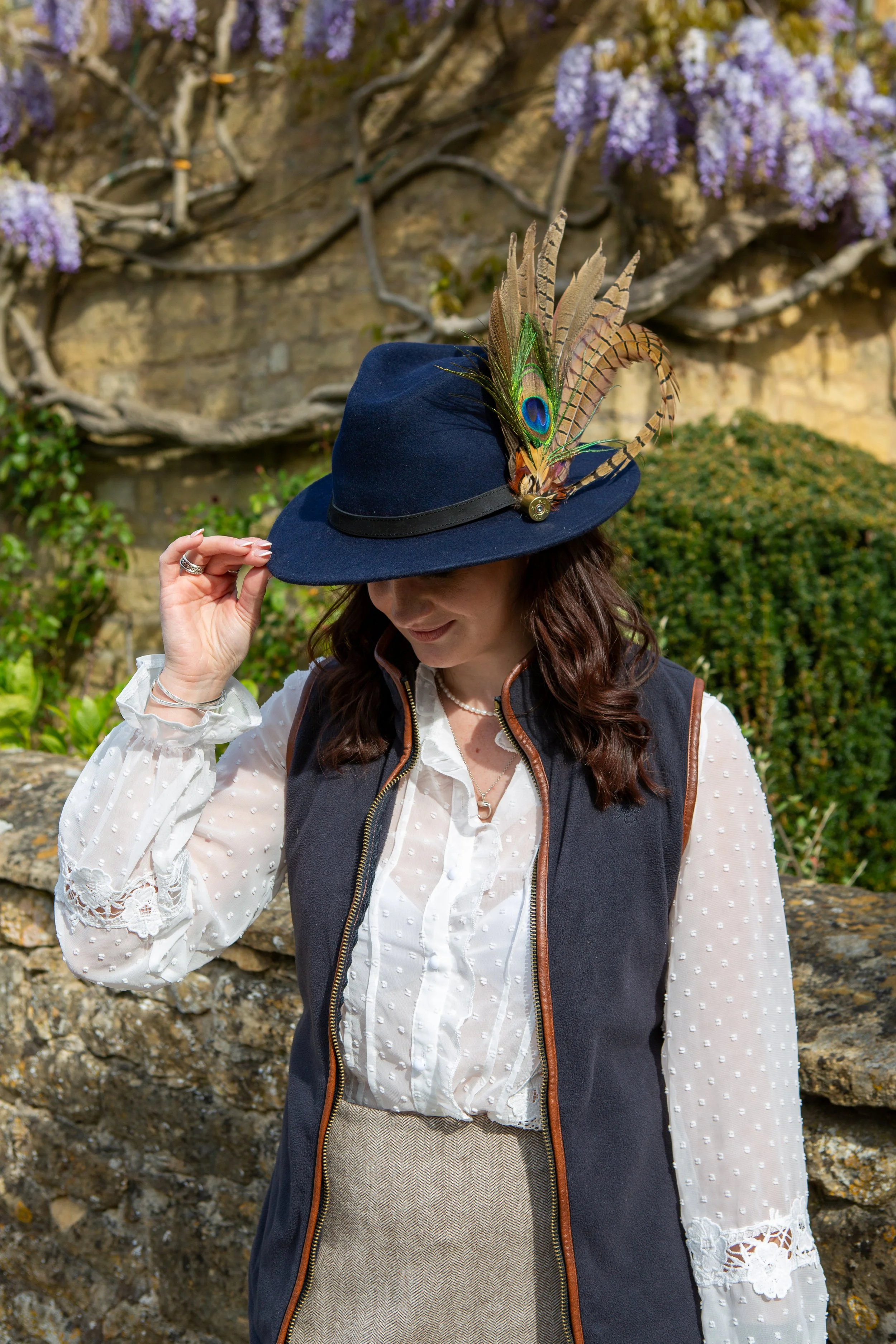 A woman wearing a navy blue hat with large peacock feathers, standing outdoors in front of a stone wall covered with purple wisteria flowers.