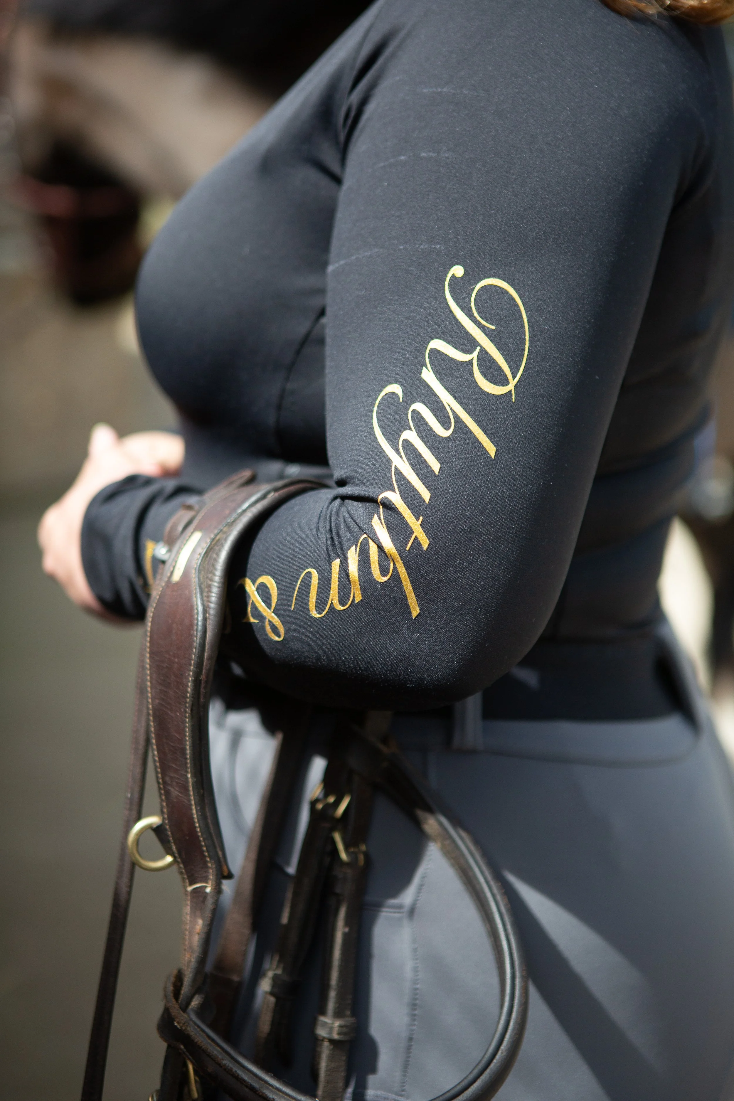 Close-up of a person wearing a dark jacket with gold lettering on the sleeve and holding a leather riding crop, with a horse in the background.