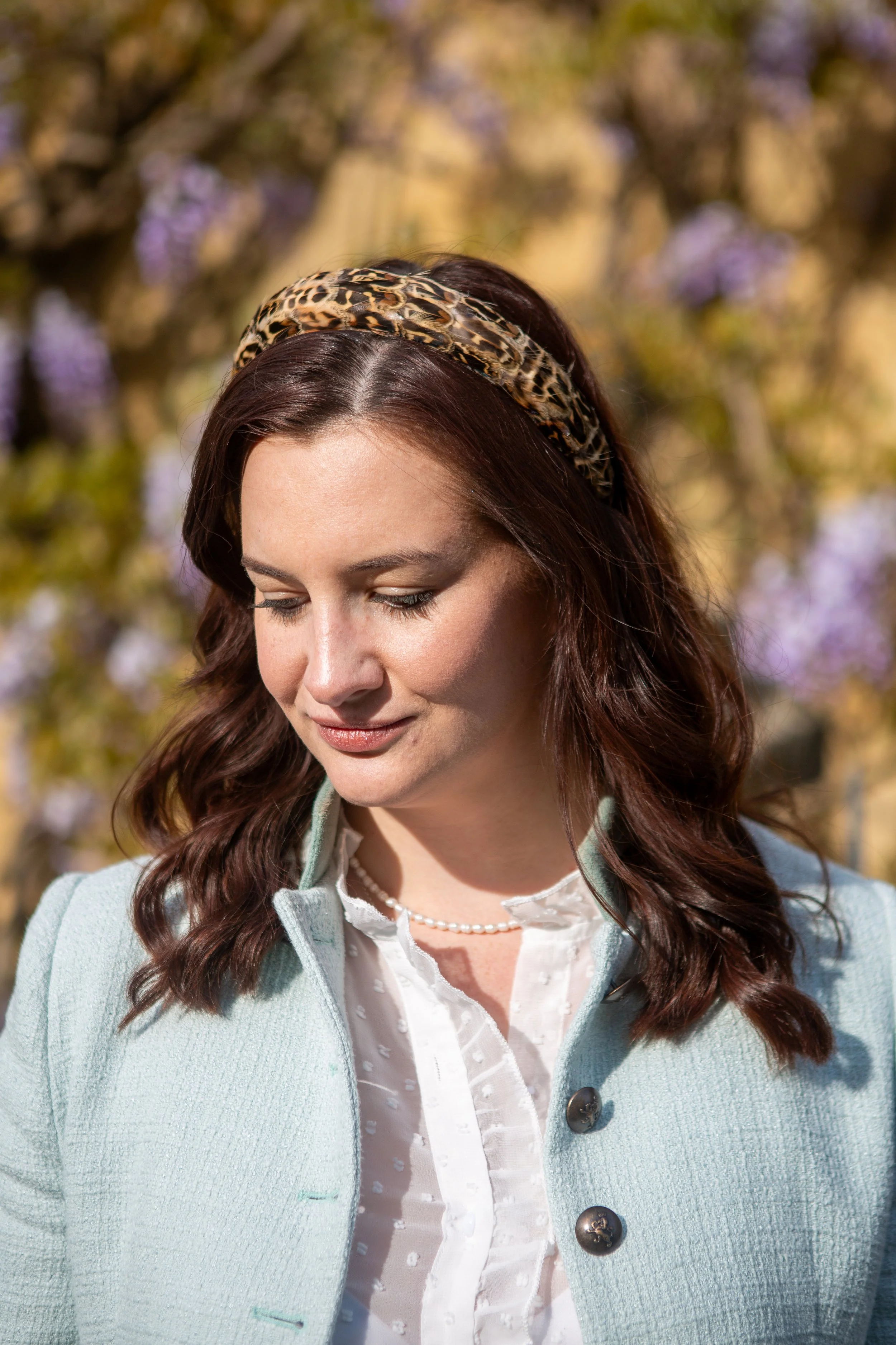 A young woman with brown hair wearing a leopard-print headband, a pearl necklace, a white blouse with textured dots, and a light blue coat with large buttons, looking down outdoors with blurred flowering trees in the background.