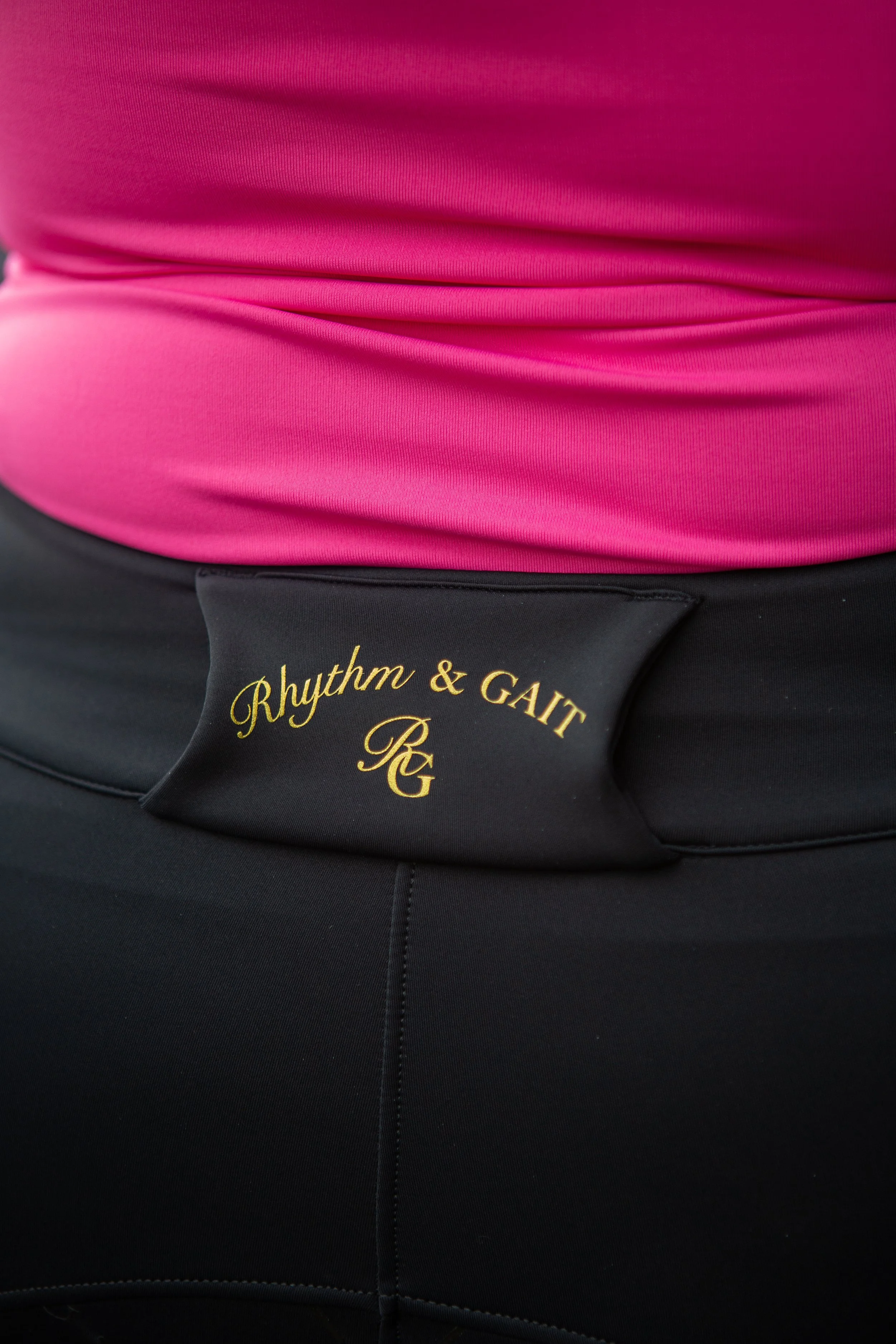 Close-up of a pink athletic top with a black band below that has gold embroidery reading "Rhythm & Gait".