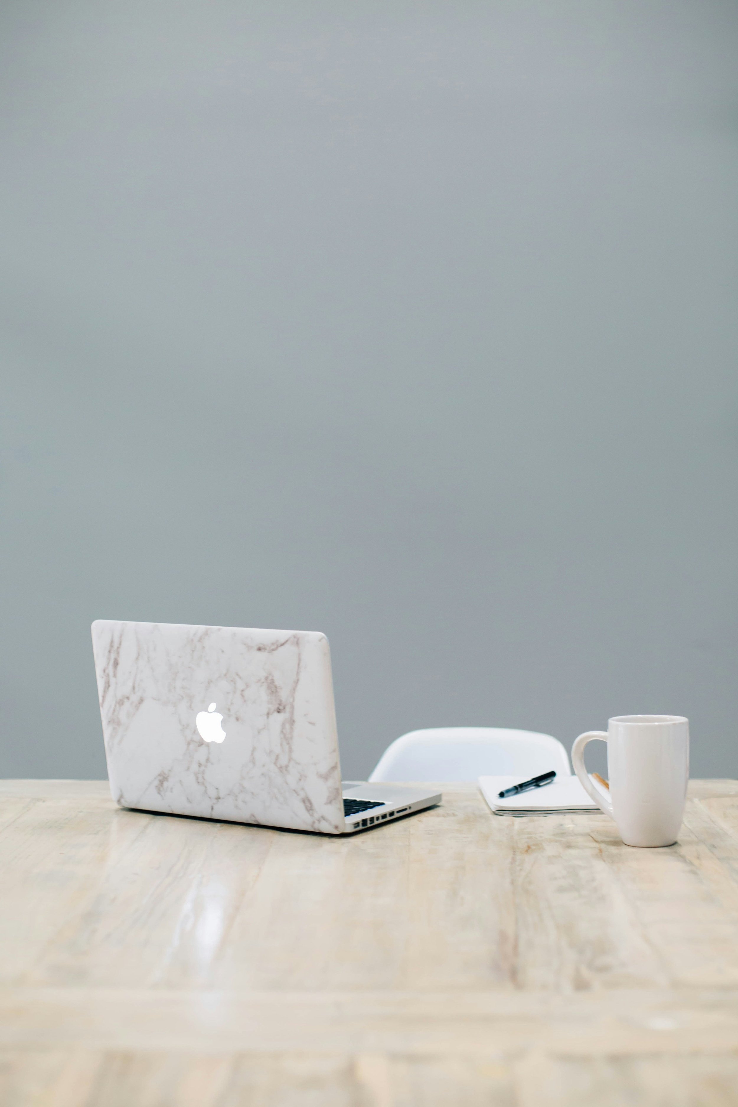 A marble laptop on a wooden table, with a white coffee mug, a black pen, and a notepad or tablet nearby, in front of a gray wall.