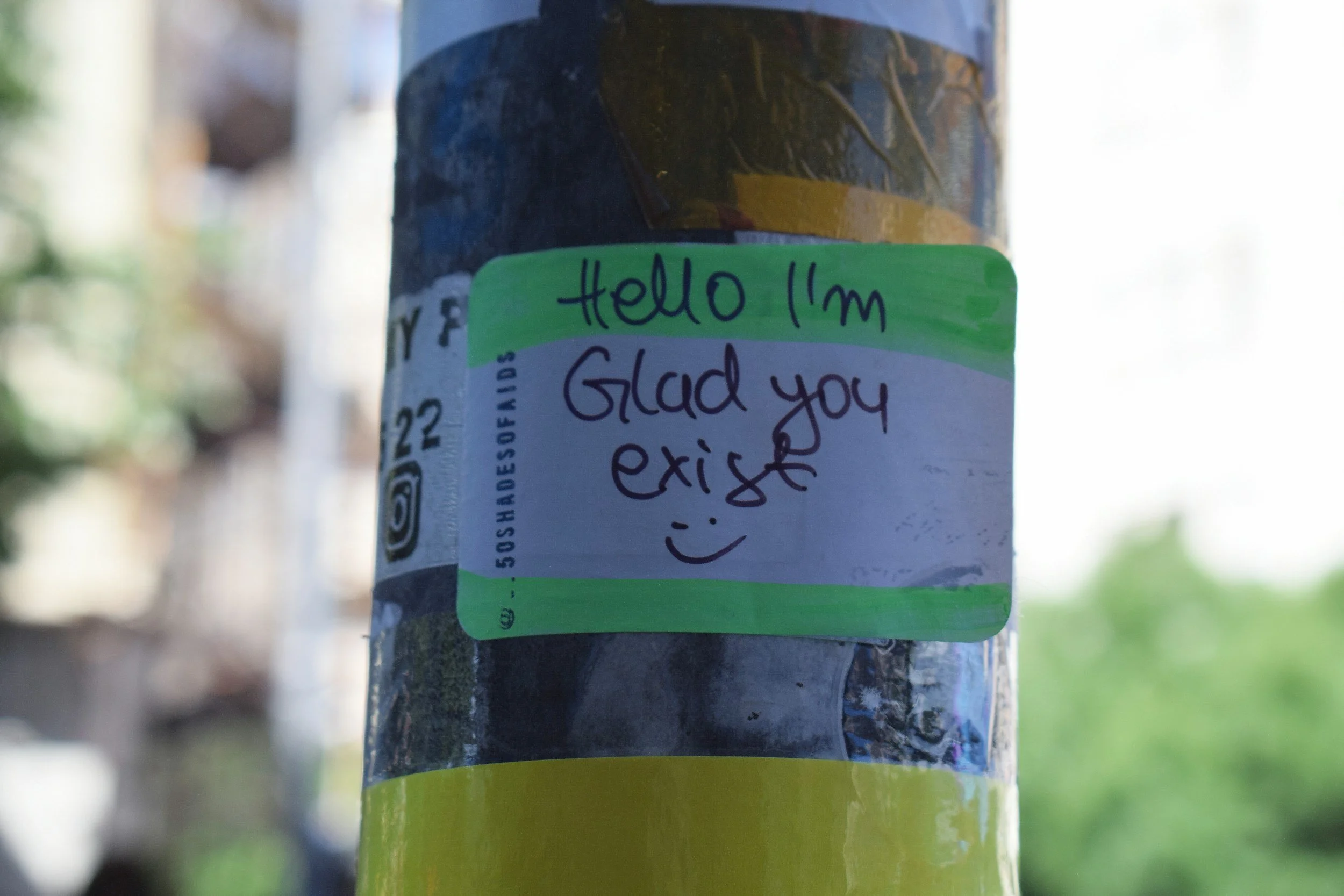 A close-up of a lamppost with a green sticky note that says "Hello I’m Glad you exist" handwritten on it. The lamppost is outdoors with a blurred background of nature.