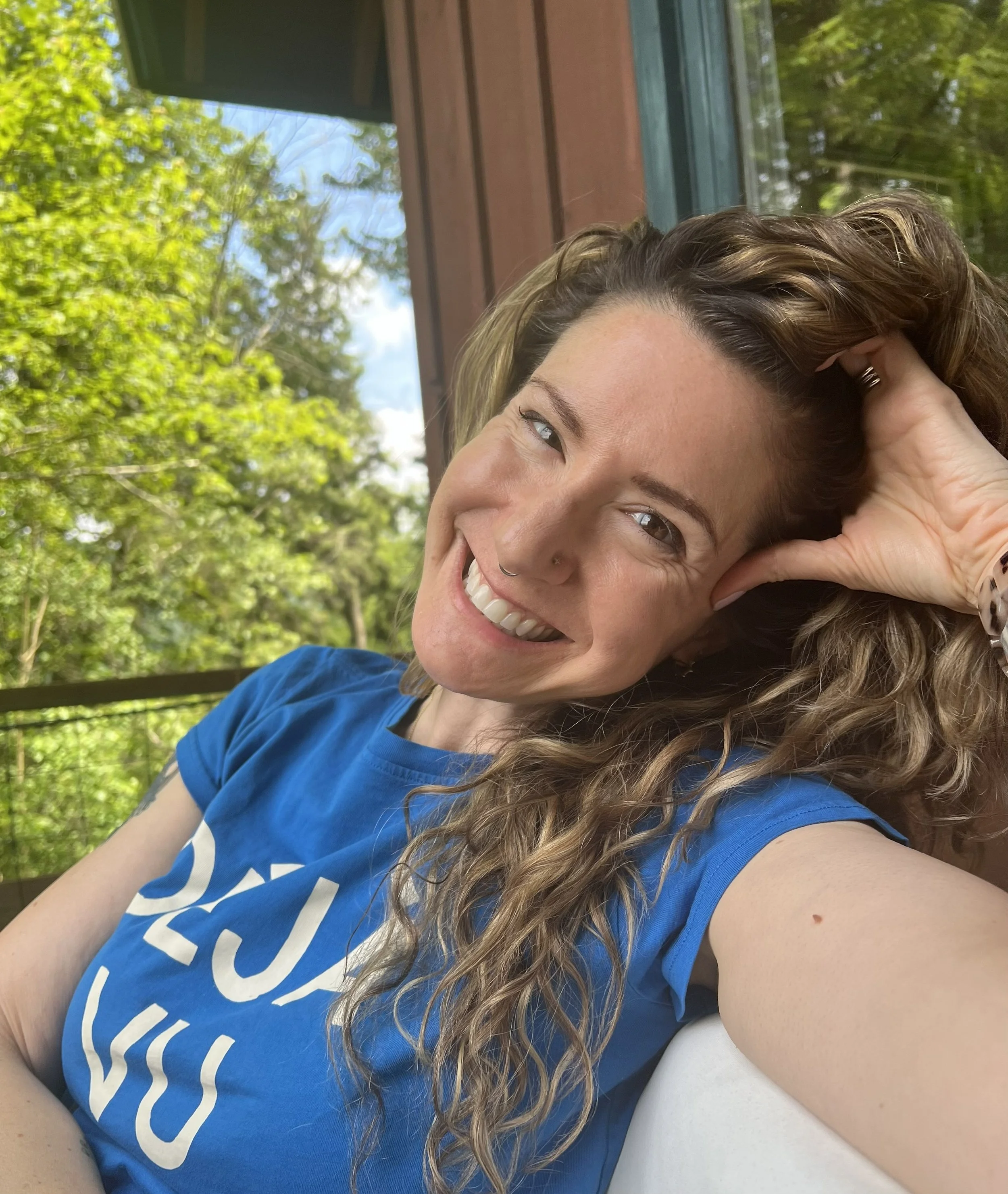 Close-up of a smiling woman with long curly hair, sitting outdoors with greenery in the background, wearing a blue T-shirt.