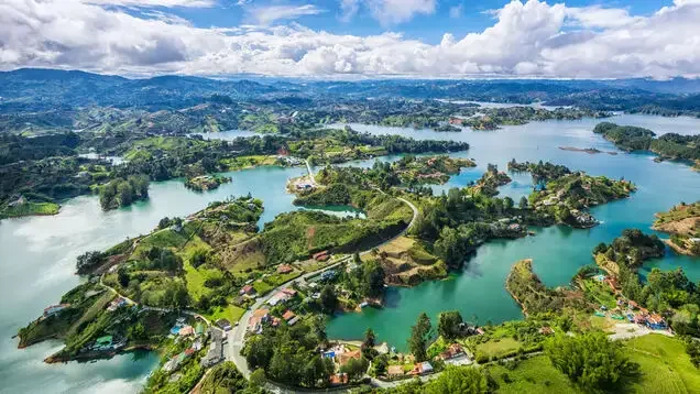 Aerial view of a landscape with lakes, green hills, trees, and scattered houses under a partly cloudy sky.